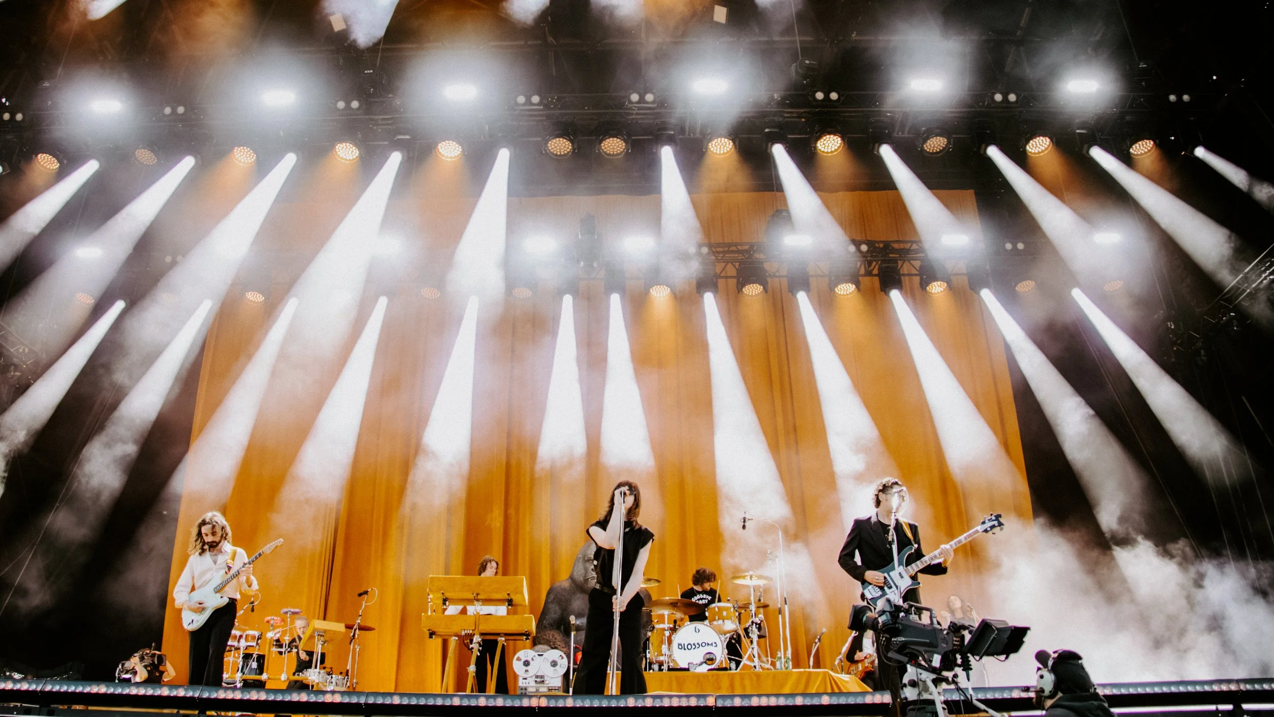A band performs on stage with bright stage lights and fog effects, with musicians playing guitars, drums, and a microphone, in a concert setting.