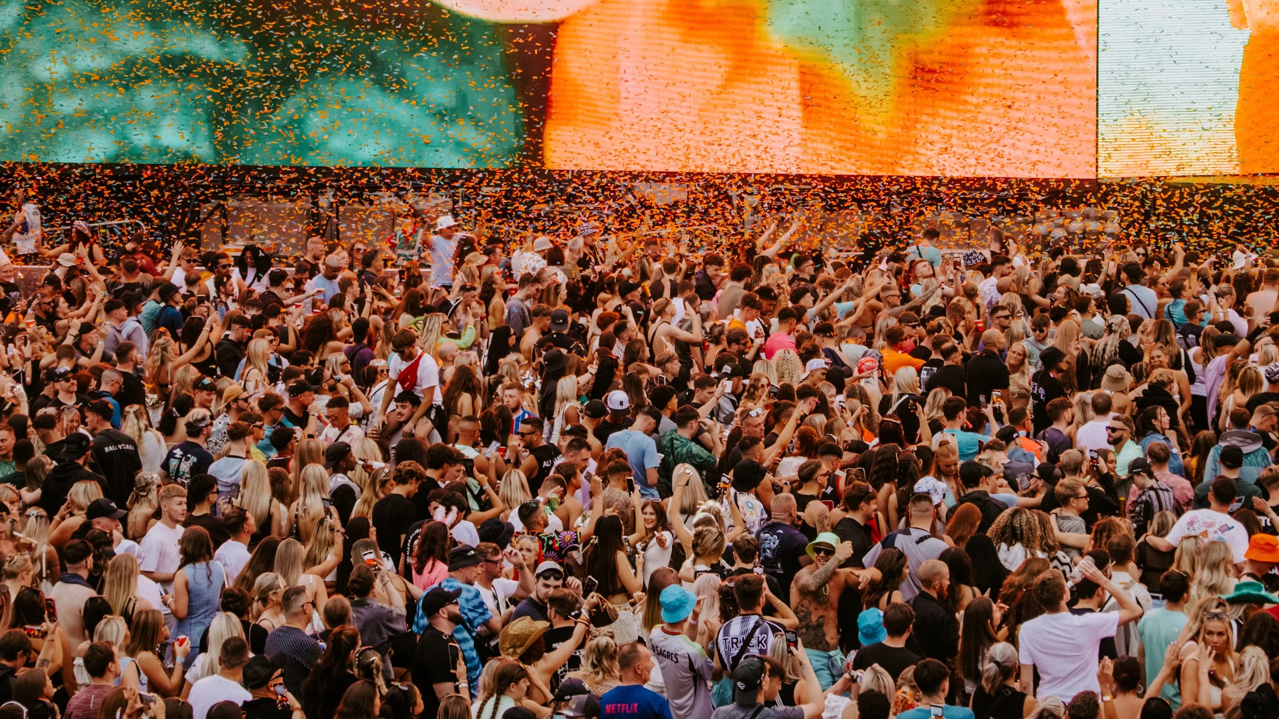 A large crowd of people attending an outdoor concert or festival, with orange confetti falling from the sky and a large screen in the background.