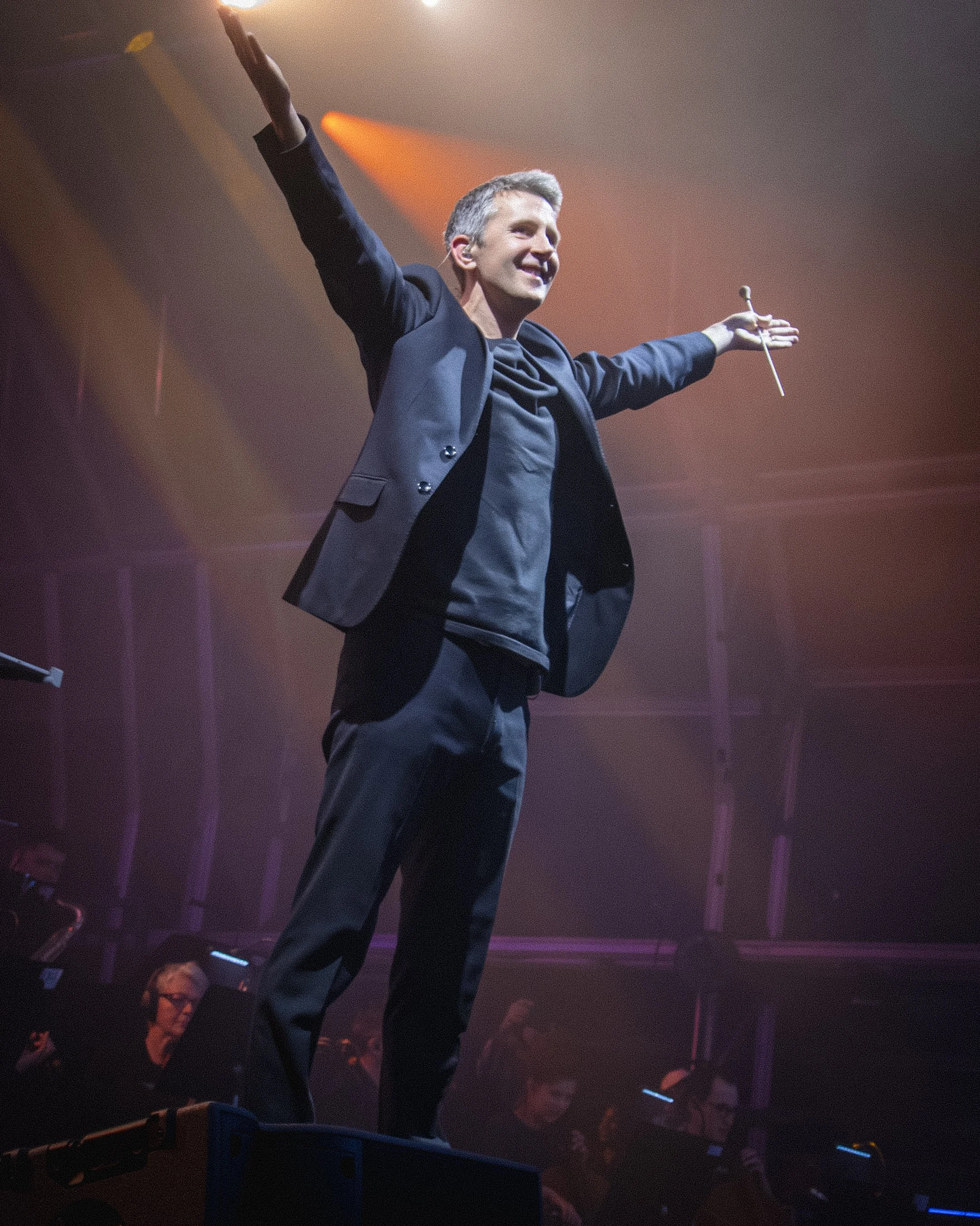 A man in a suit with outstretched arms holding a conductor's baton is smiling on stage with a warm spotlight. The audience and a woman with glasses are visible in the background.