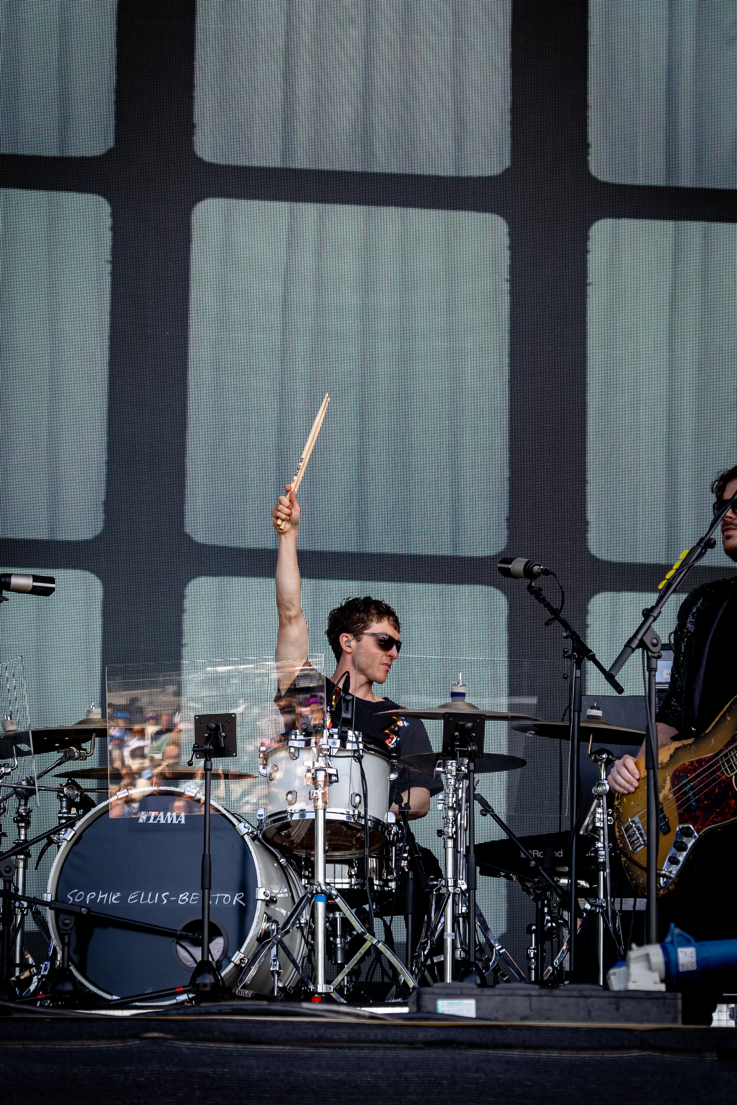 A drummer with short hair and sunglasses playing on stage during a music concert, with a large electronic screen in the background showing black grid patterns. The drum set has a bass drum labeled 'Sophie Elis-Betor'. A hand with a drumstick is raise