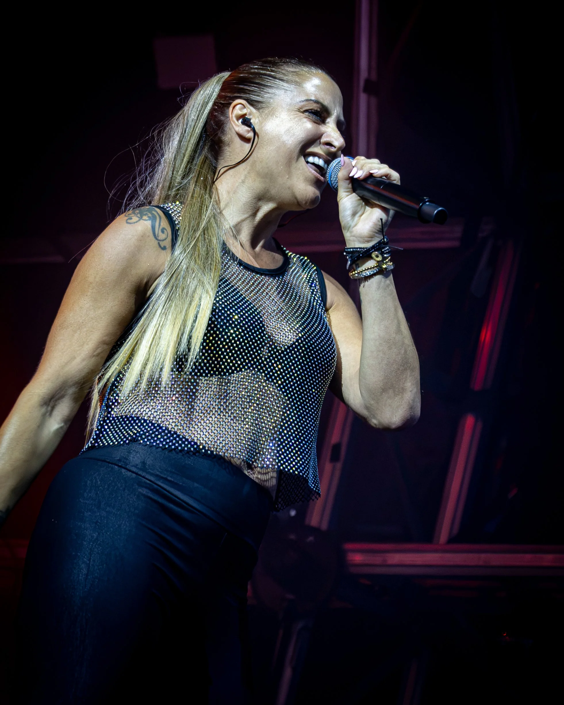 A female singer performs on stage, holding a microphone close to her mouth, wearing a sleeveless, metallic, mesh top, with long blonde hair, and wearing a black earpiece.