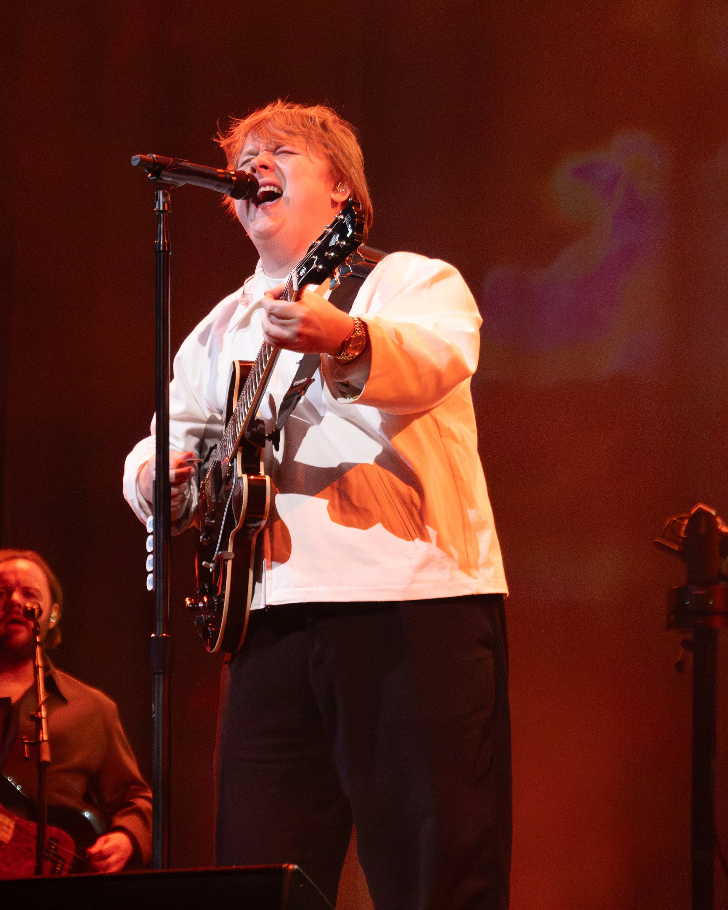 A person with short hair singing into a microphone and playing an electric guitar on stage, with two women in the background, one holding a guitar and the other standing beside her.