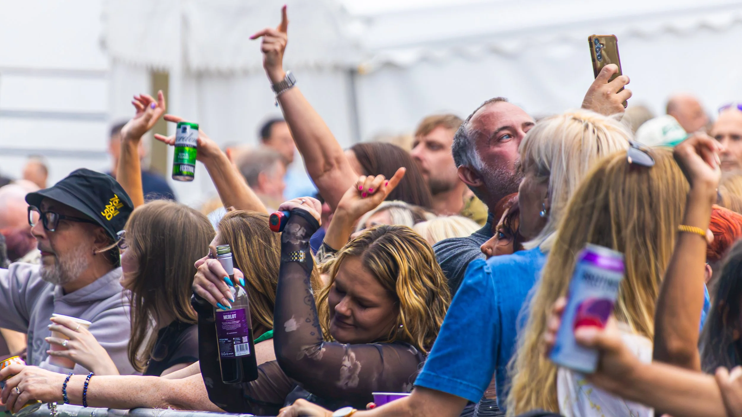 Crowd of people at an outdoor event, some holding drinks, raising hands, and taking photos.
