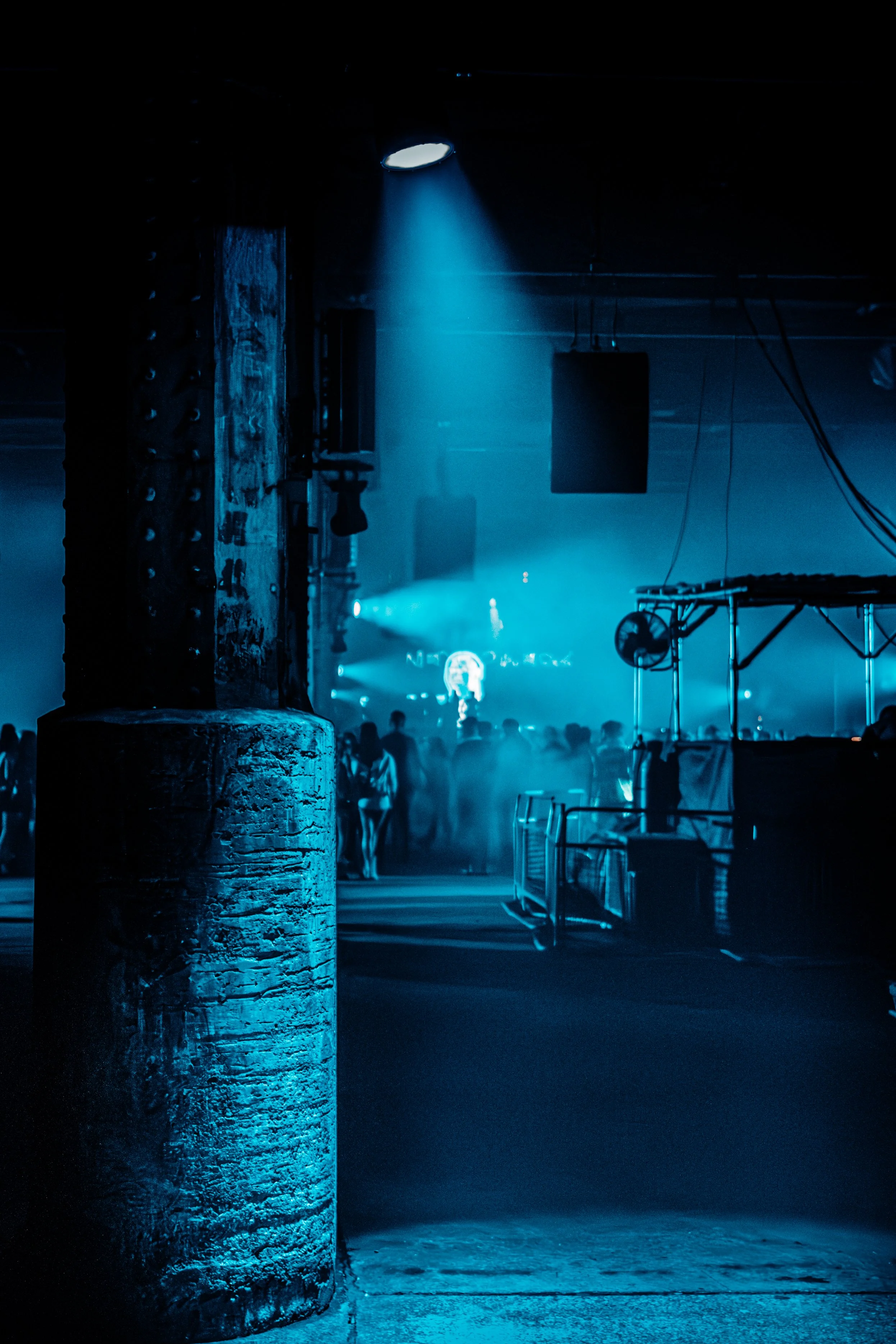 Dimly lit nightclub scene with a spotlight, smoky atmosphere, crowd of people, and stage equipment.