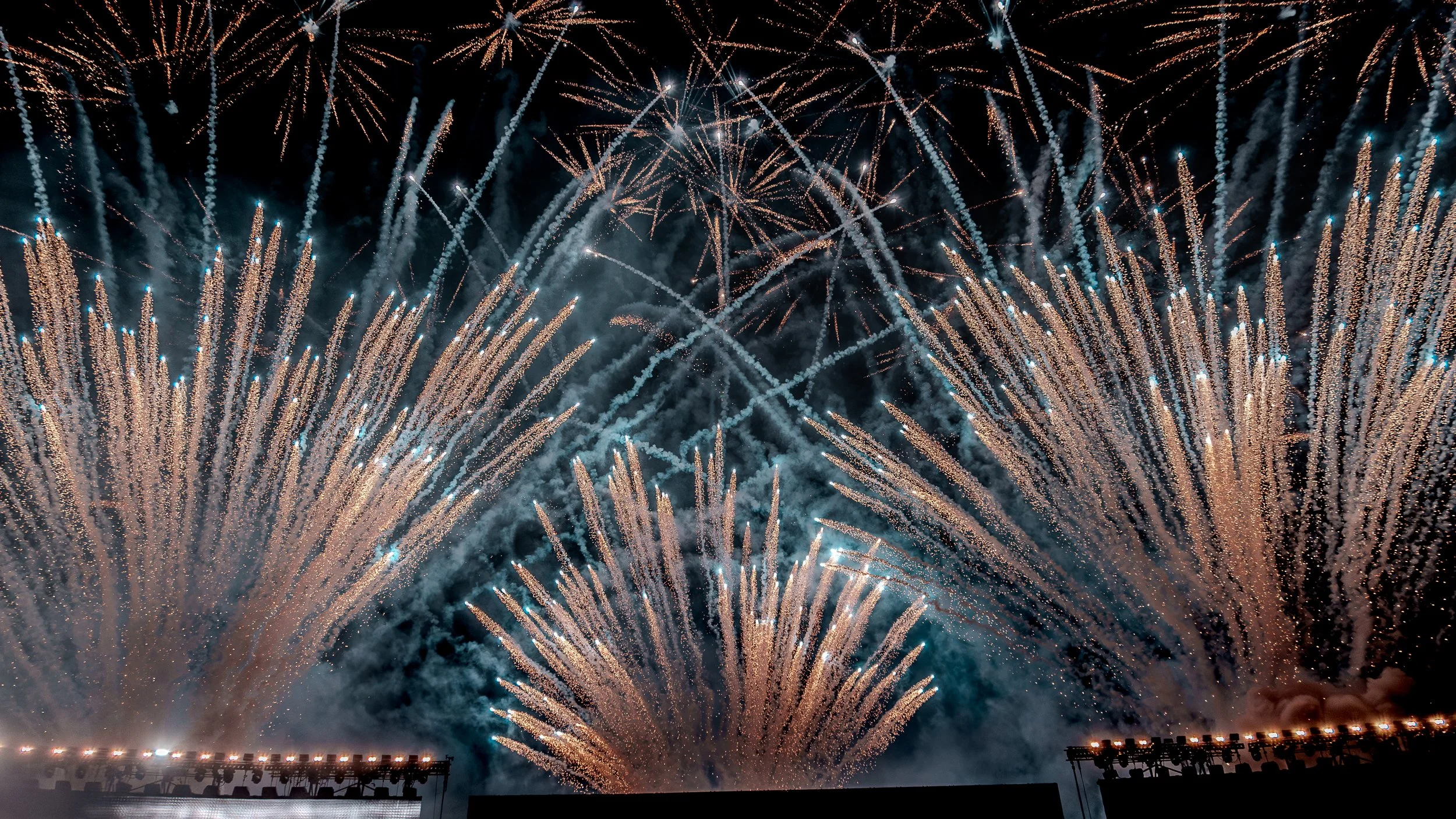 Spectacular fireworks display lighting up the night sky with bursts of gold, white, and blue. Smoke and trails from explosions are visible, with a dark silhouette of a stadium or stage in the foreground.