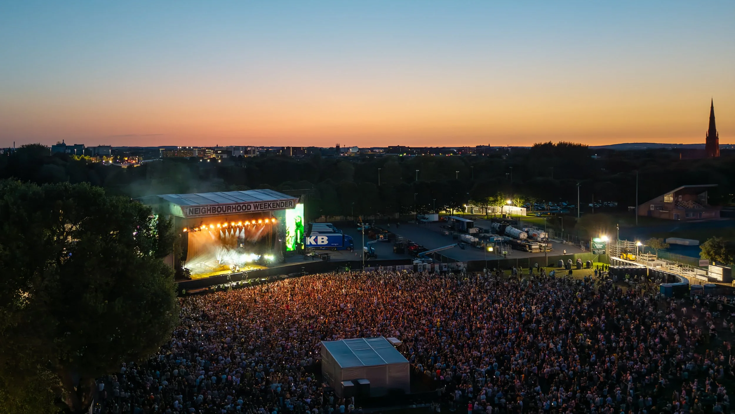An outdoor concert during sunset with a large crowd facing a brightly lit stage, and a cityscape with buildings and a church steeple in the background.