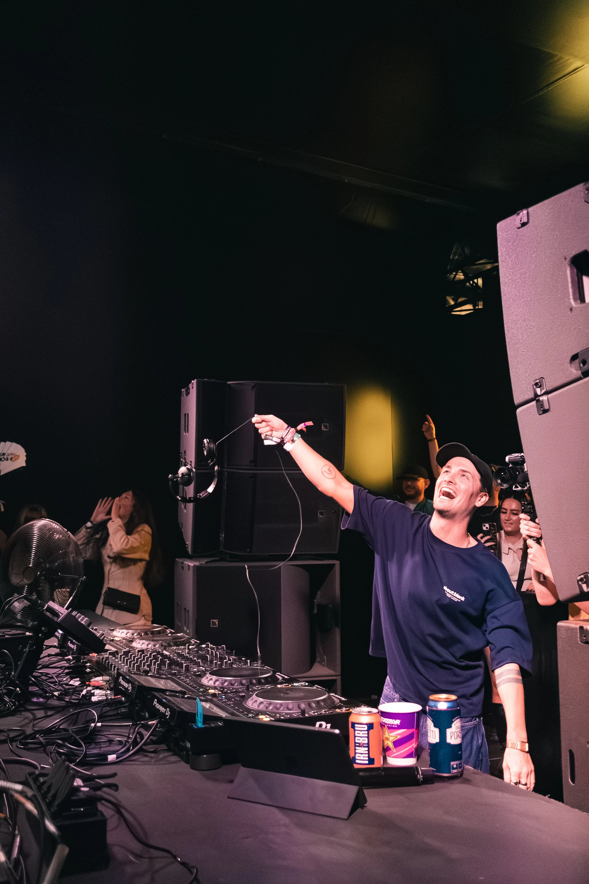 A DJ and a person with a raised arm at a party or concert, with sound equipment and beverages on the table, in a dark indoor setting with audience members visible.
