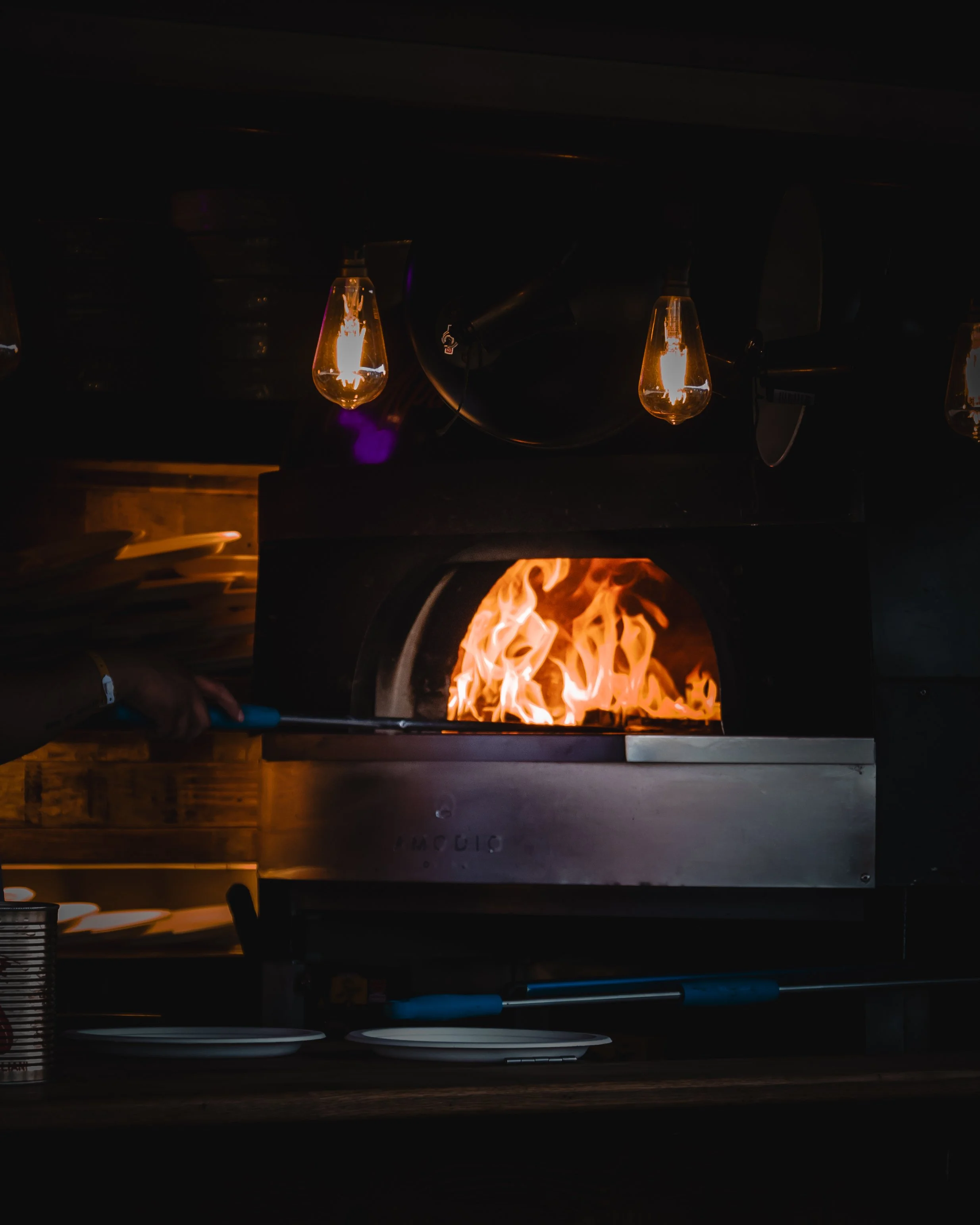 An open pizza oven with flames inside, a pizza being cooked, and a stove with glowing lights above.