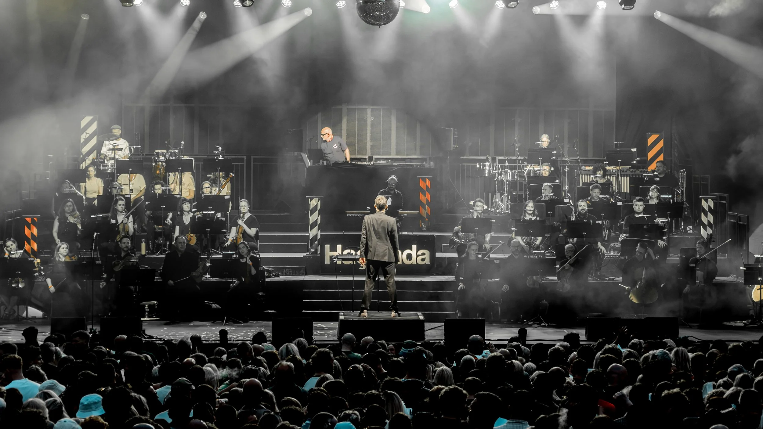 A large orchestra on stage performing at a concert, with a conductor in the foreground and multiple musicians playing instruments, illuminated by bright stage lights and surrounded by fog.