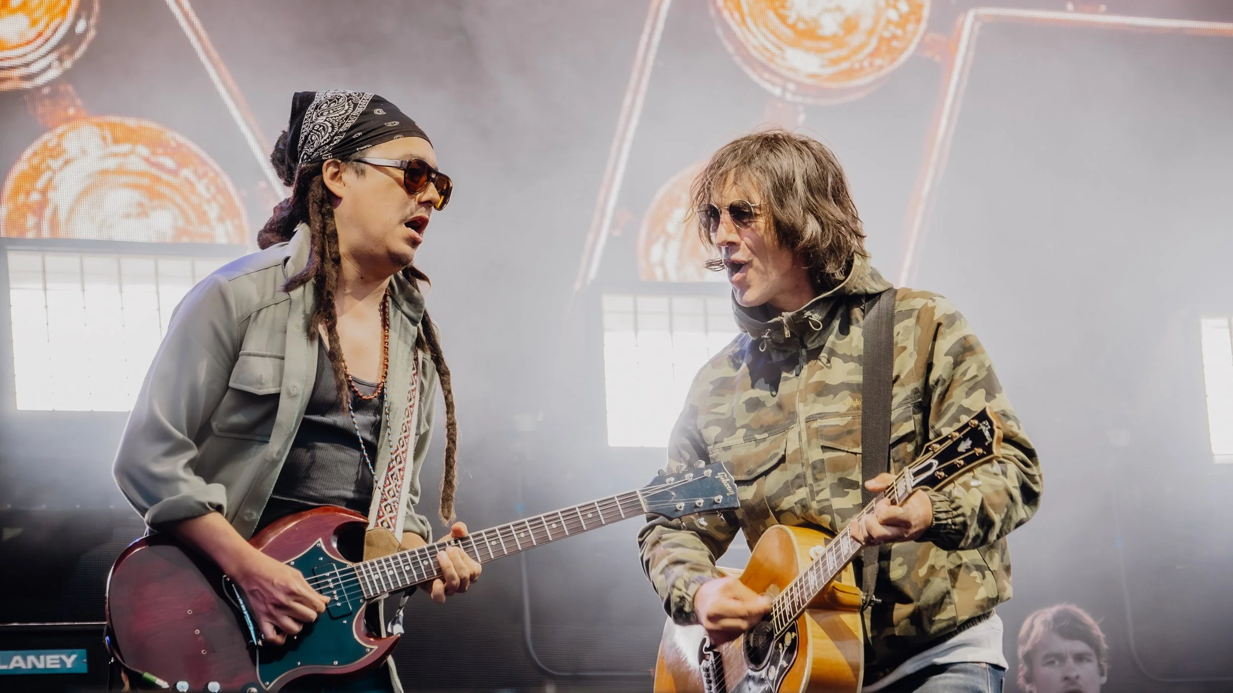 Two musicians with guitars singing together on stage, wearing sunglasses, with a bright background of hanging lights and screens.