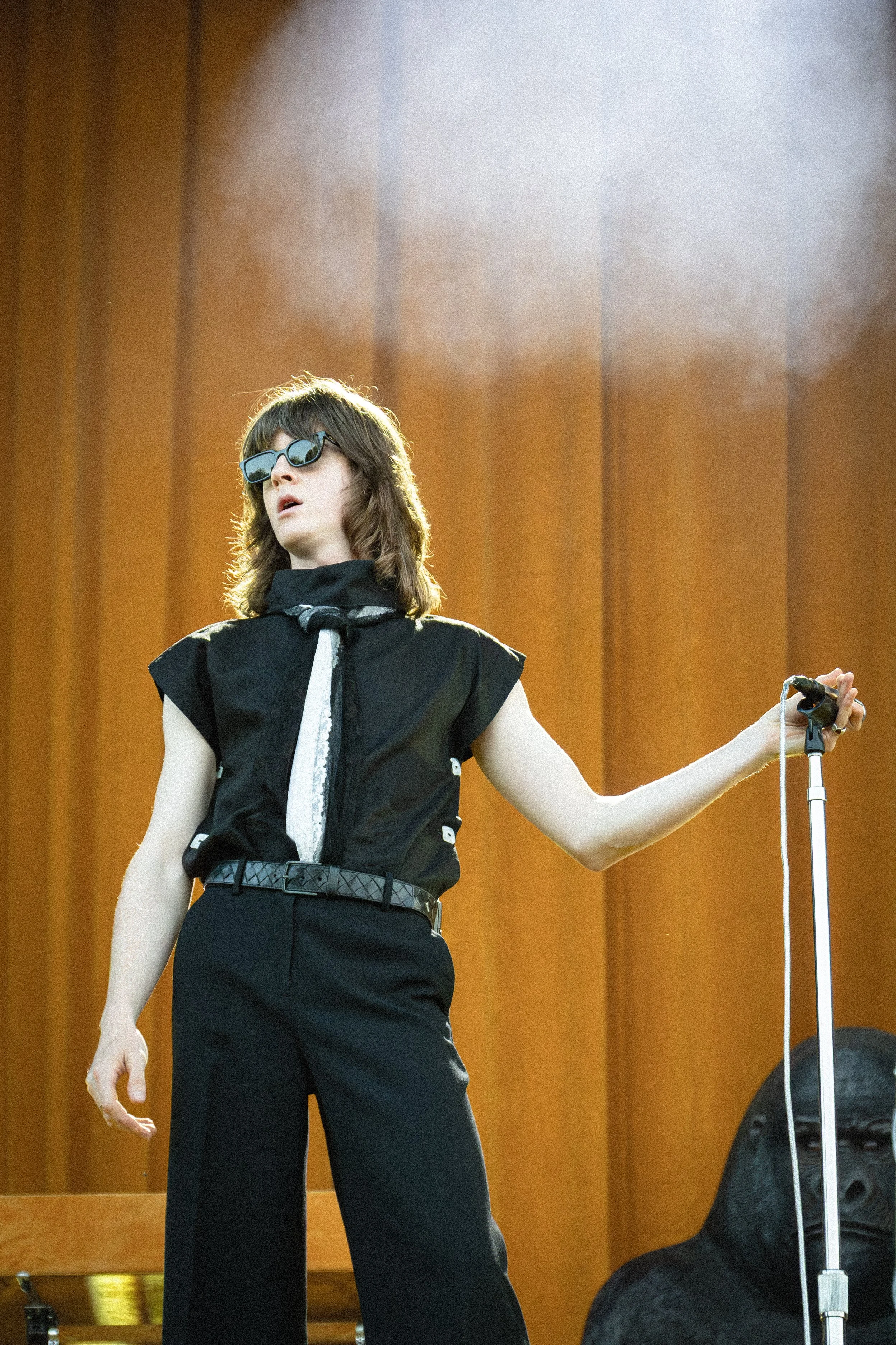 A woman in black sunglasses, a black vest, and black pants standing in front of a wooden wall, holding a microphone with a black dog lying on the floor nearby.
