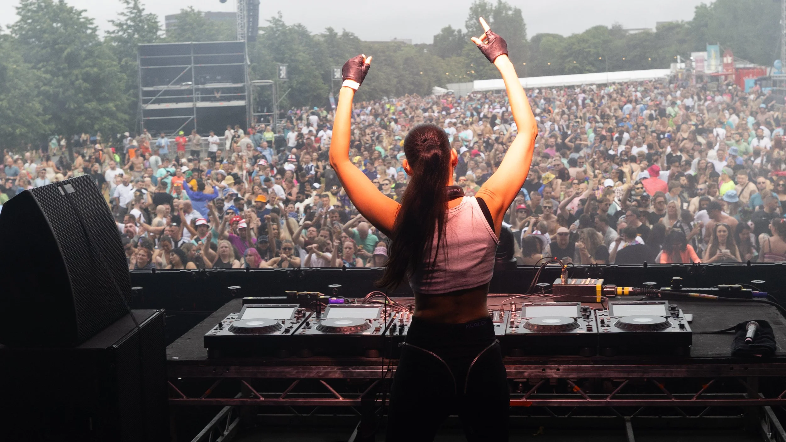 A female DJ performing on stage at an outdoor music festival with a large crowd of attendees. The DJ has her arms raised and is facing the audience, with mixer equipment in front of her and large speakers on either side. The crowd is enjoying the mus