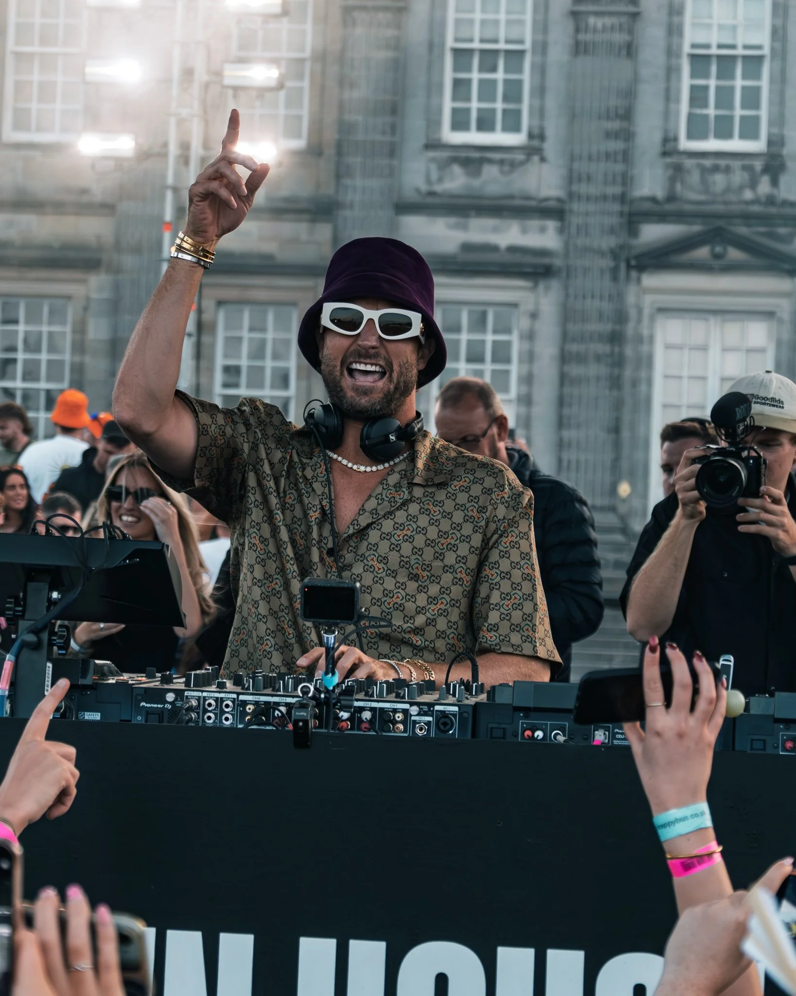 A man wearing sunglasses, a purple hat, and a patterned shirt spins DJ controls at an outdoor event, surrounded by a crowd with some holding smartphones.