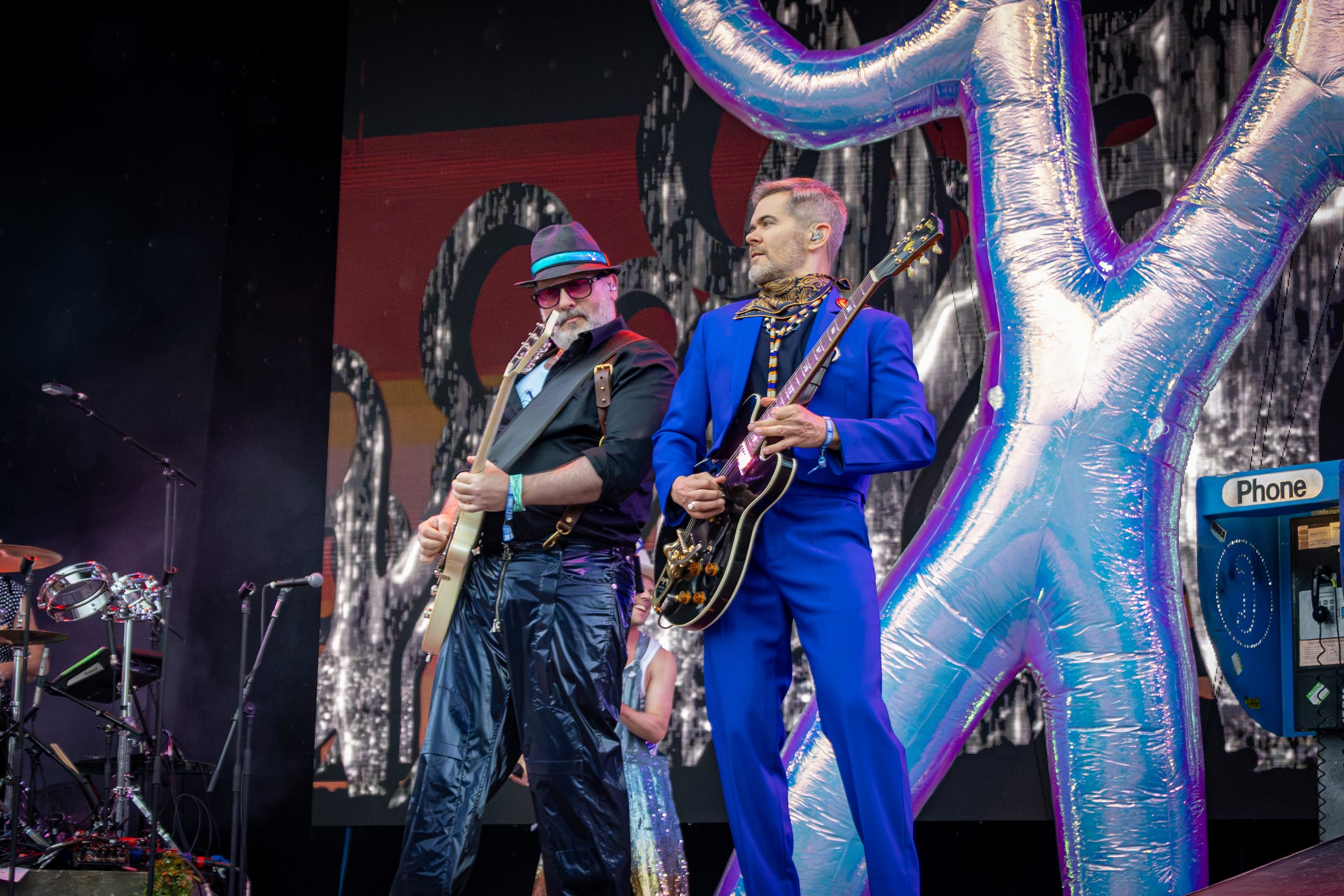 Two male musicians performing on stage, one wearing a fedora, sunglasses, and a dark shirt, and the other in a vibrant blue suit with a patterned scarf around his neck. They are playing guitars, with a background of black and red visuals and large si