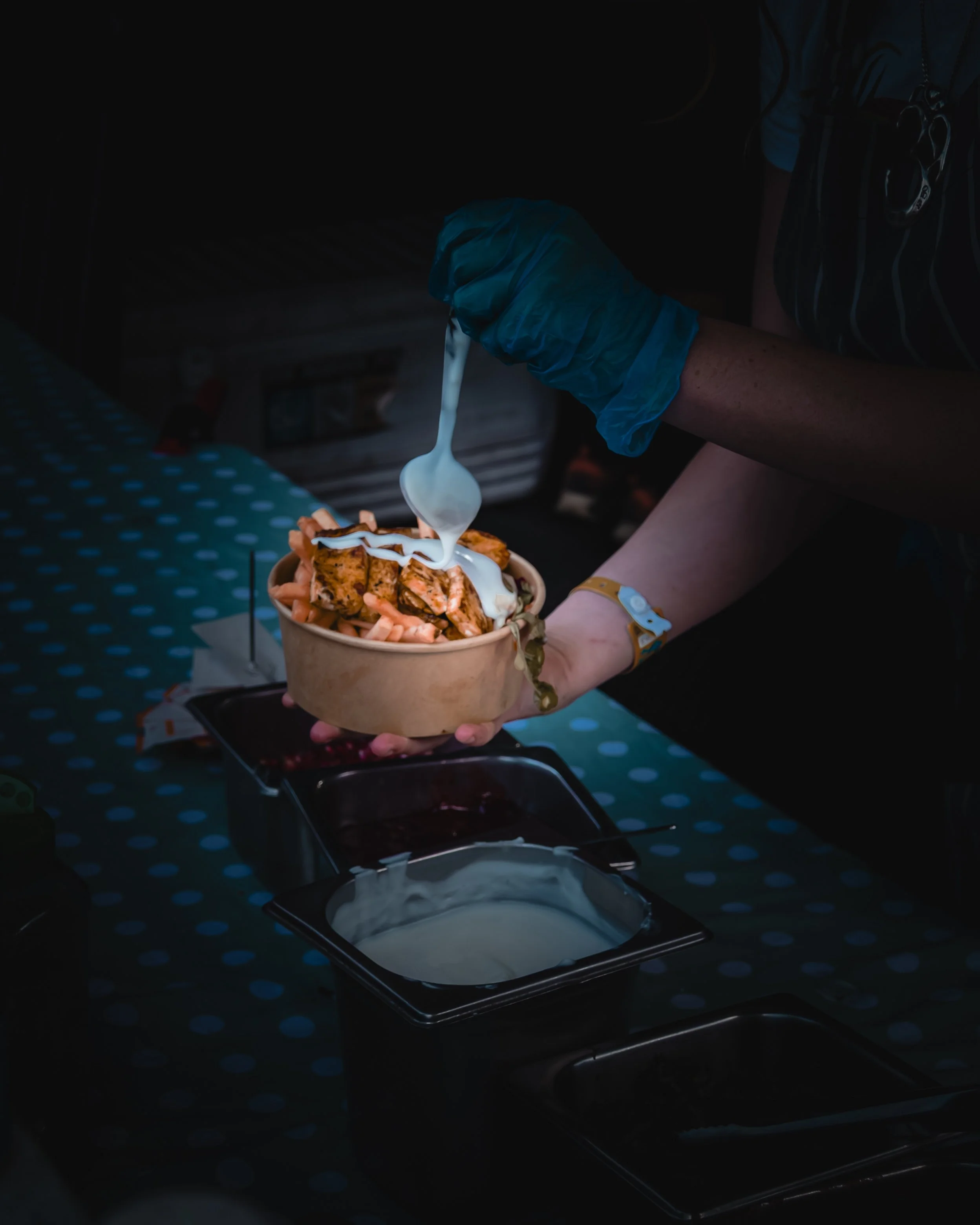 Person wearing a blue glove adding a swirl of white sauce to a bowl of fries topped with chicken and gravy, with a dark background and a black device at the bottom.