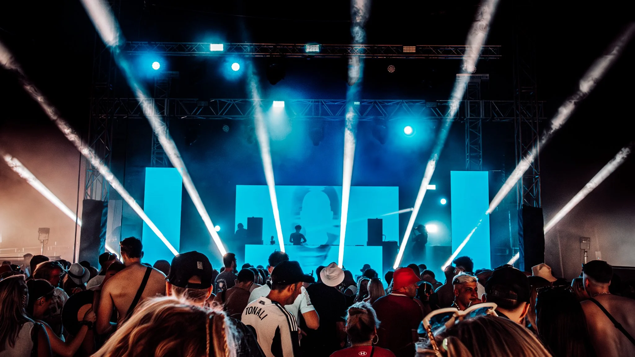 A large crowd at an outdoor music concert with a stage illuminated by bright blue and white lights, featuring performers and a DJ booth, with a night sky background.
