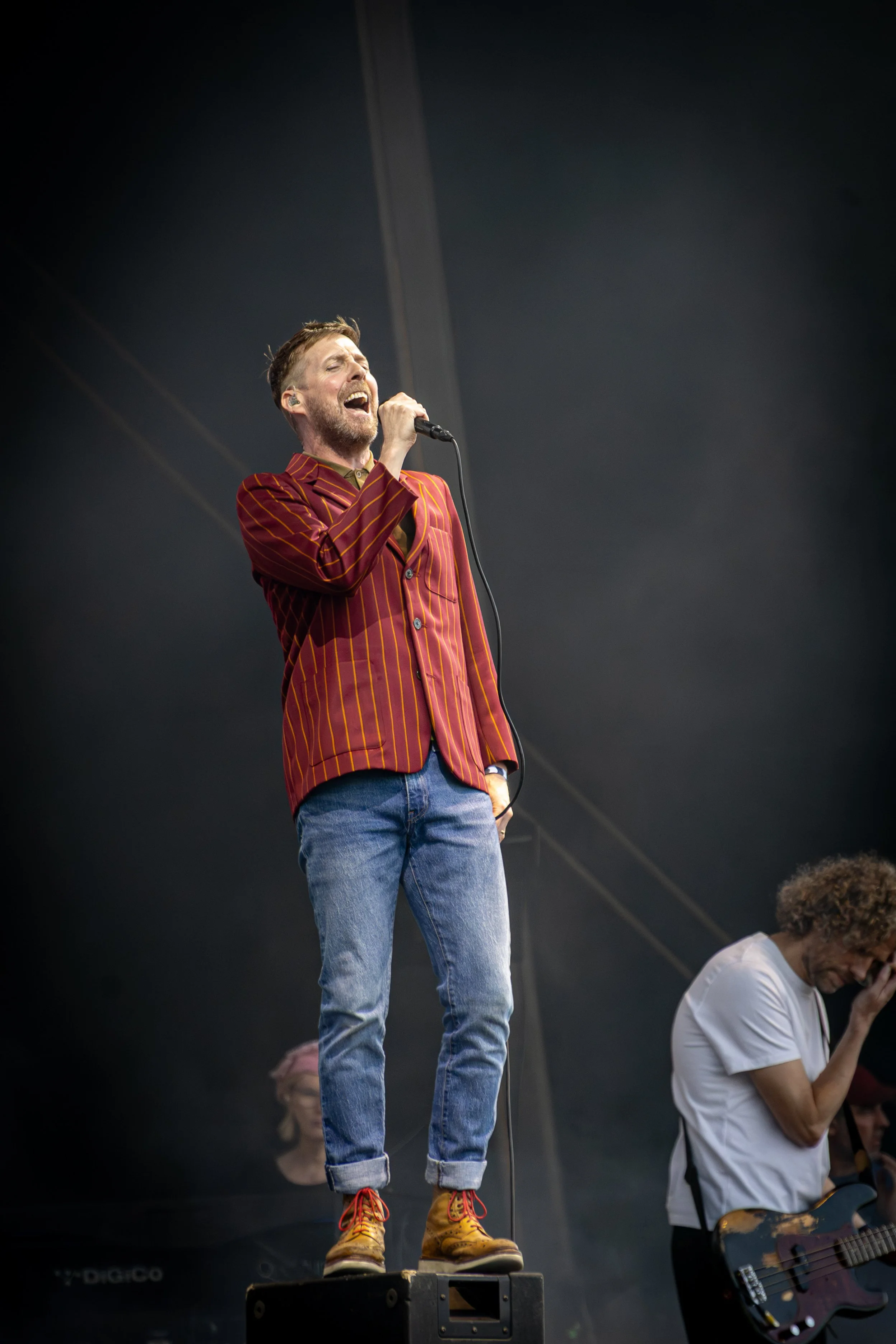 A man singing into a microphone on stage, wearing a red and yellow striped blazer, blue jeans, and yellow boots, with a dark background.