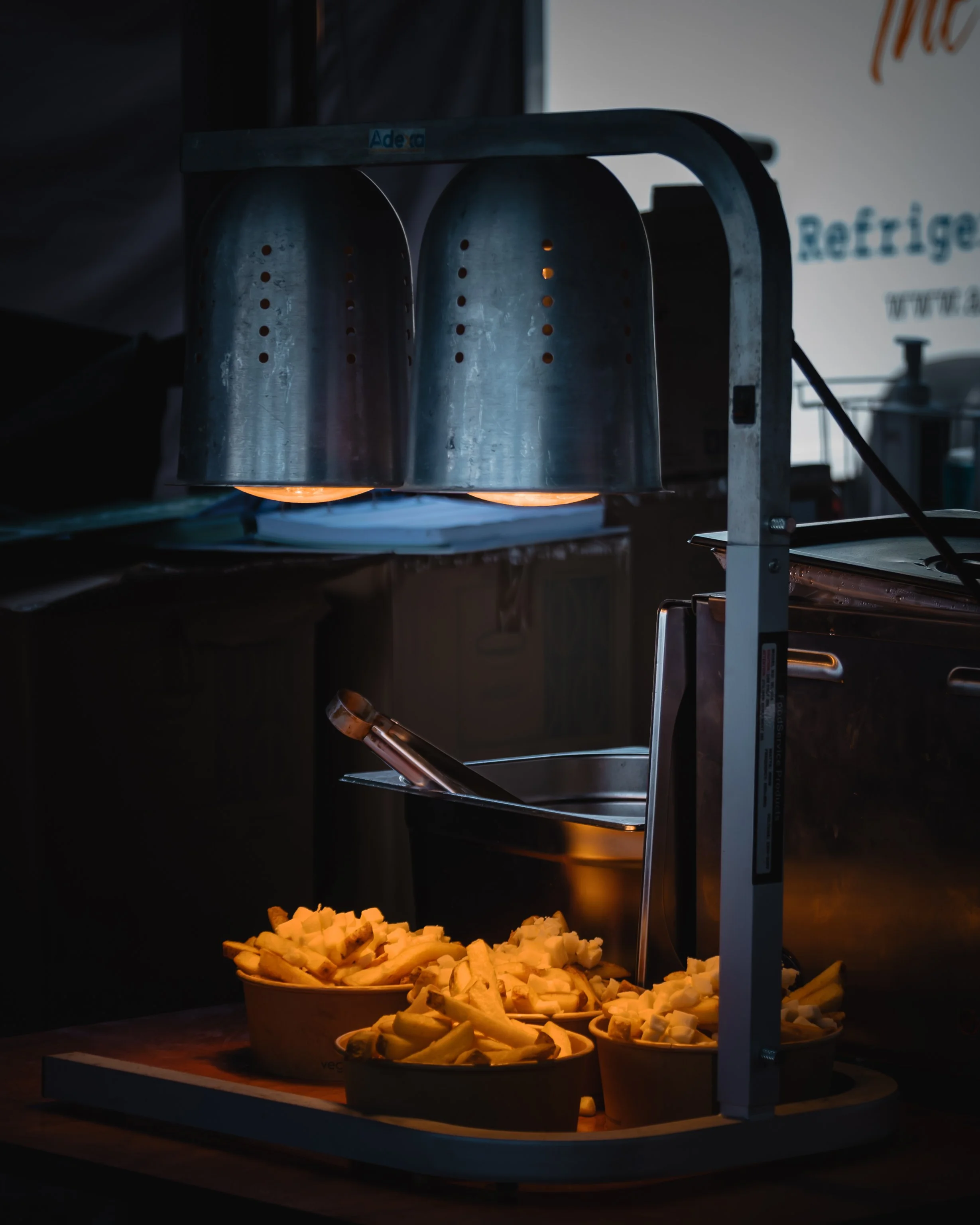 Slices of cheese or food in bowls below a heated lamp or food warmer in a dimly lit setting.