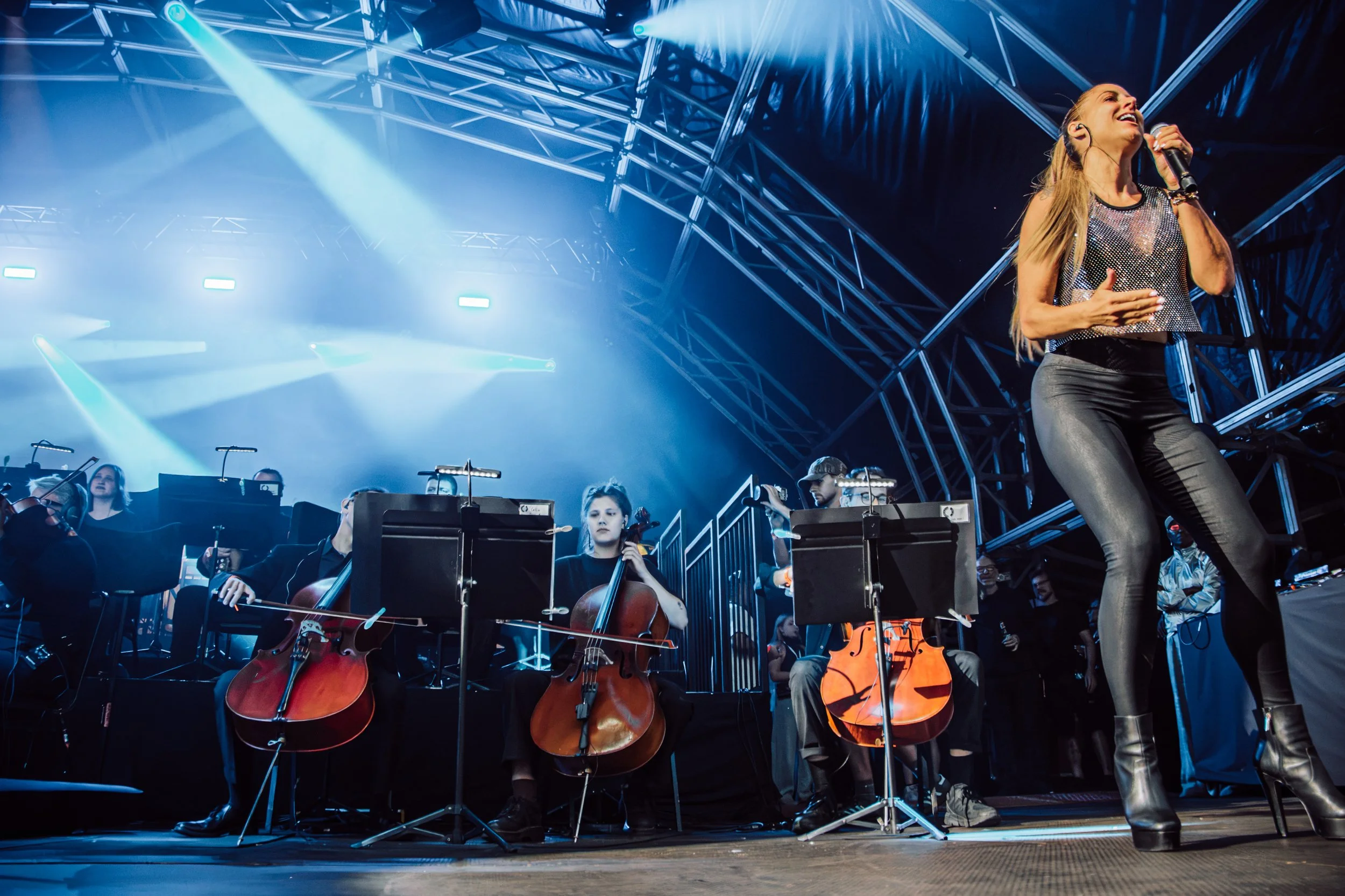 A live performance stage with a female singer holding a microphone and a large group of orchestra musicians playing string instruments, illuminated by stage lights and a metal truss structure overhead.