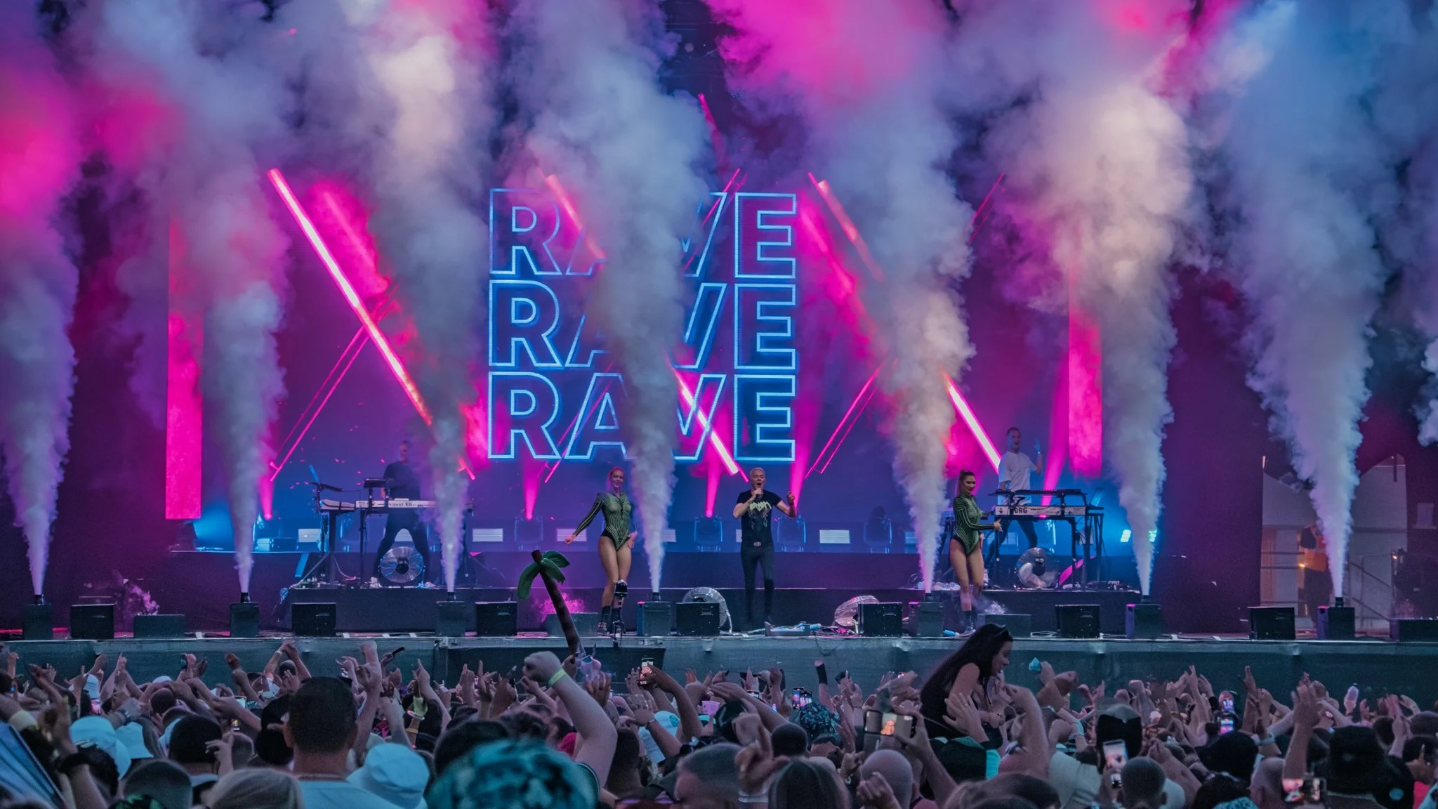 Concert stage with performers and dancers, with pink and blue neon lights and smoke effects, and large illuminated 'REVE' signs in the background.