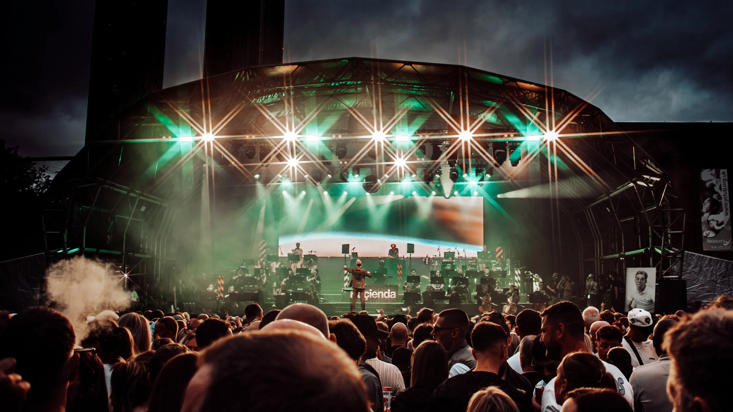 An outdoor concert with a large crowd watching a band perform on stage with vibrant green and white lighting, and a conductor in front of the orchestra.