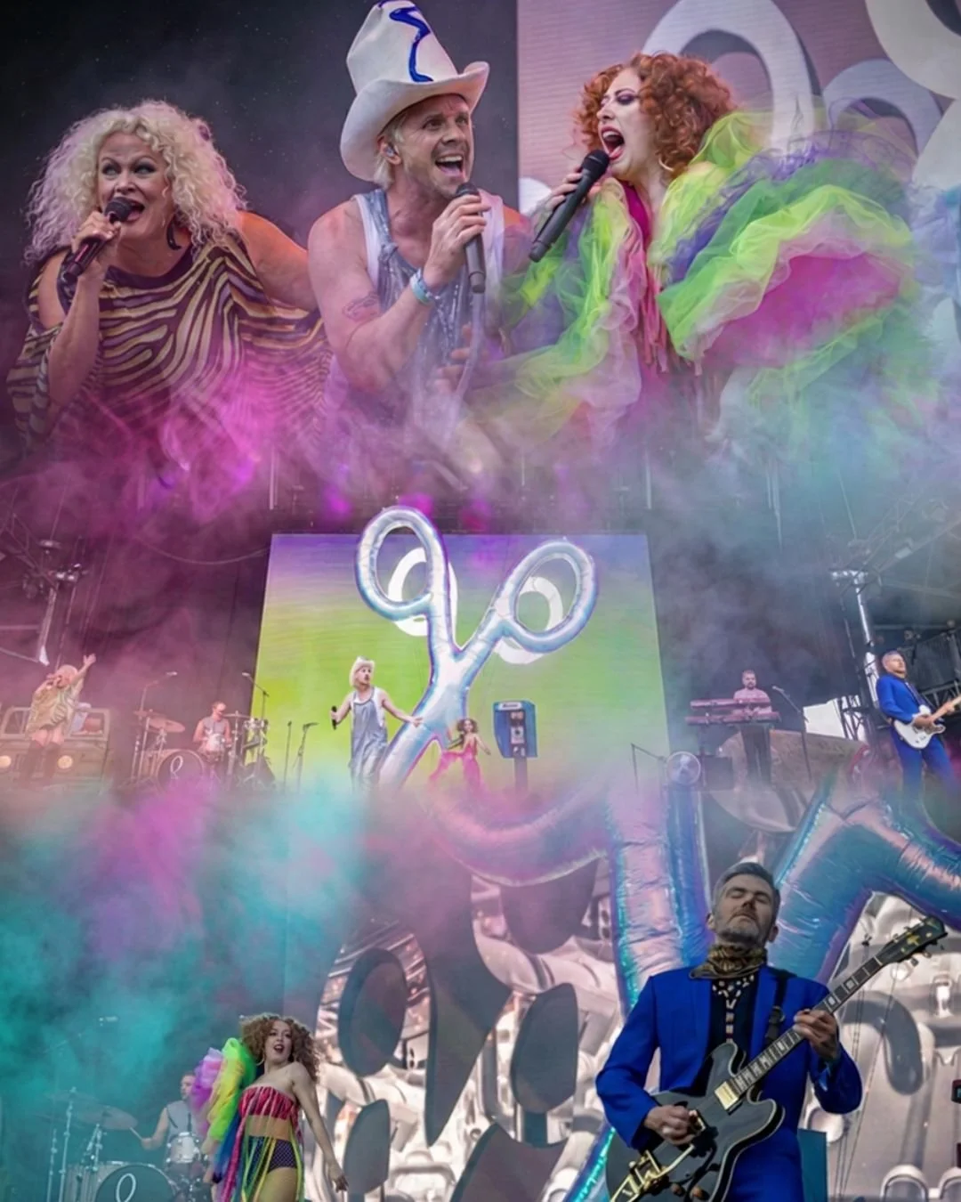 A lively stage performance featuring colorful costumes and large balloons, including a clown sculpture. Musicians and singers are performing, with smoke effects and a bright LED screen in the background.