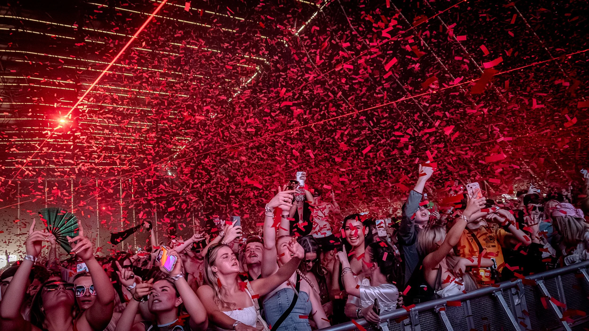 Crowd of people at a concert or event, cheering and taking photos, with red confetti falling and red lights or laser beams creating a festive atmosphere.