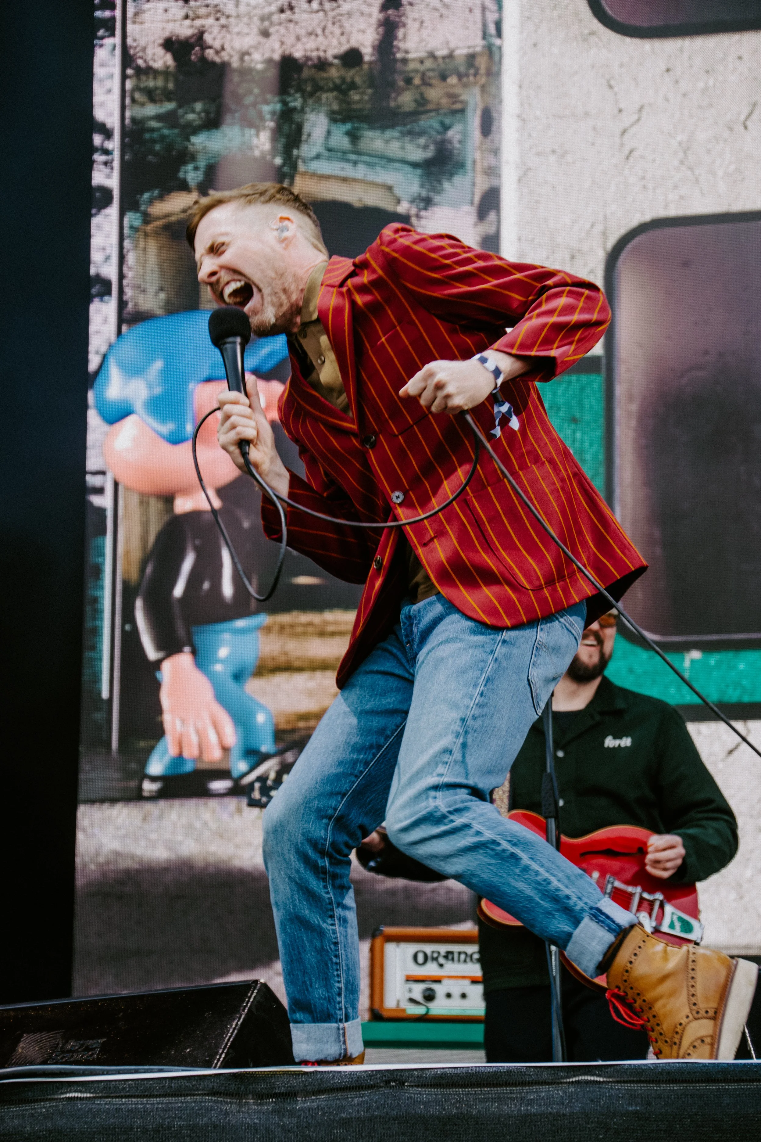 A man is singing passionately into a microphone while performing on stage, dressed in a red and yellow striped blazer, blue jeans, and tan boots, with a band in the background.