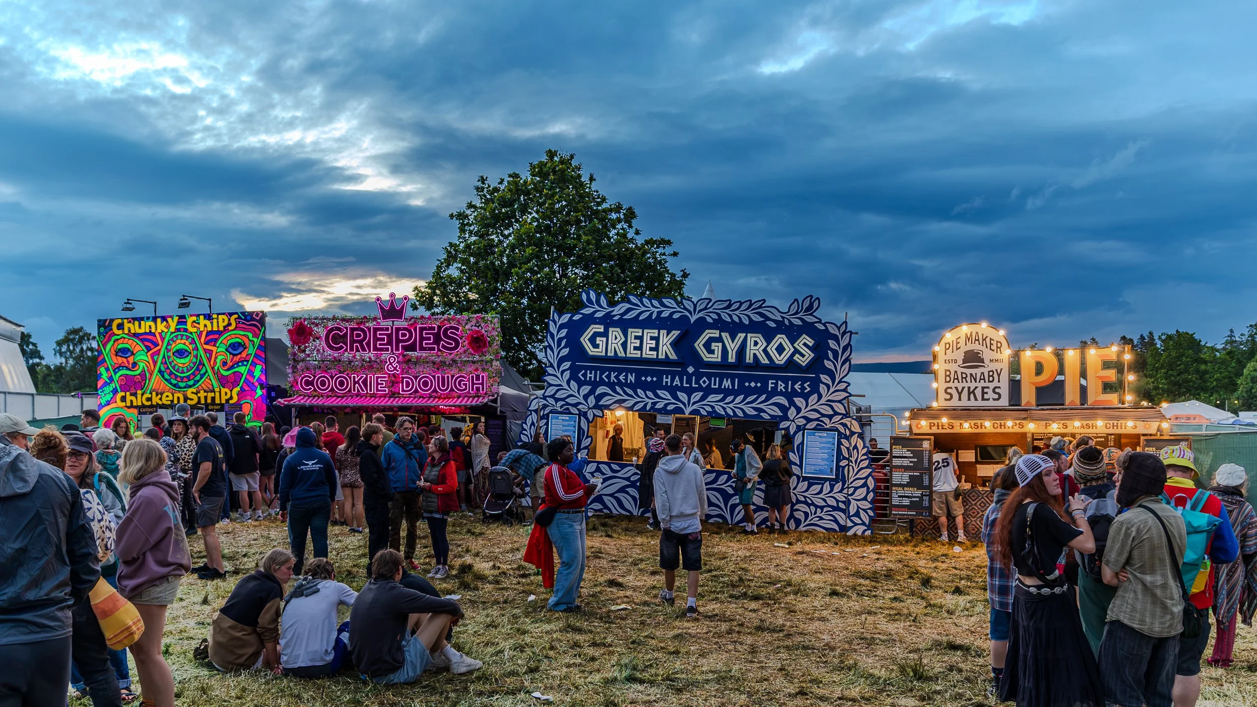 Crowd at an outdoor fair with food stalls including chicken strips, crepes, Greek gyros, and pie, under a cloudy sky.