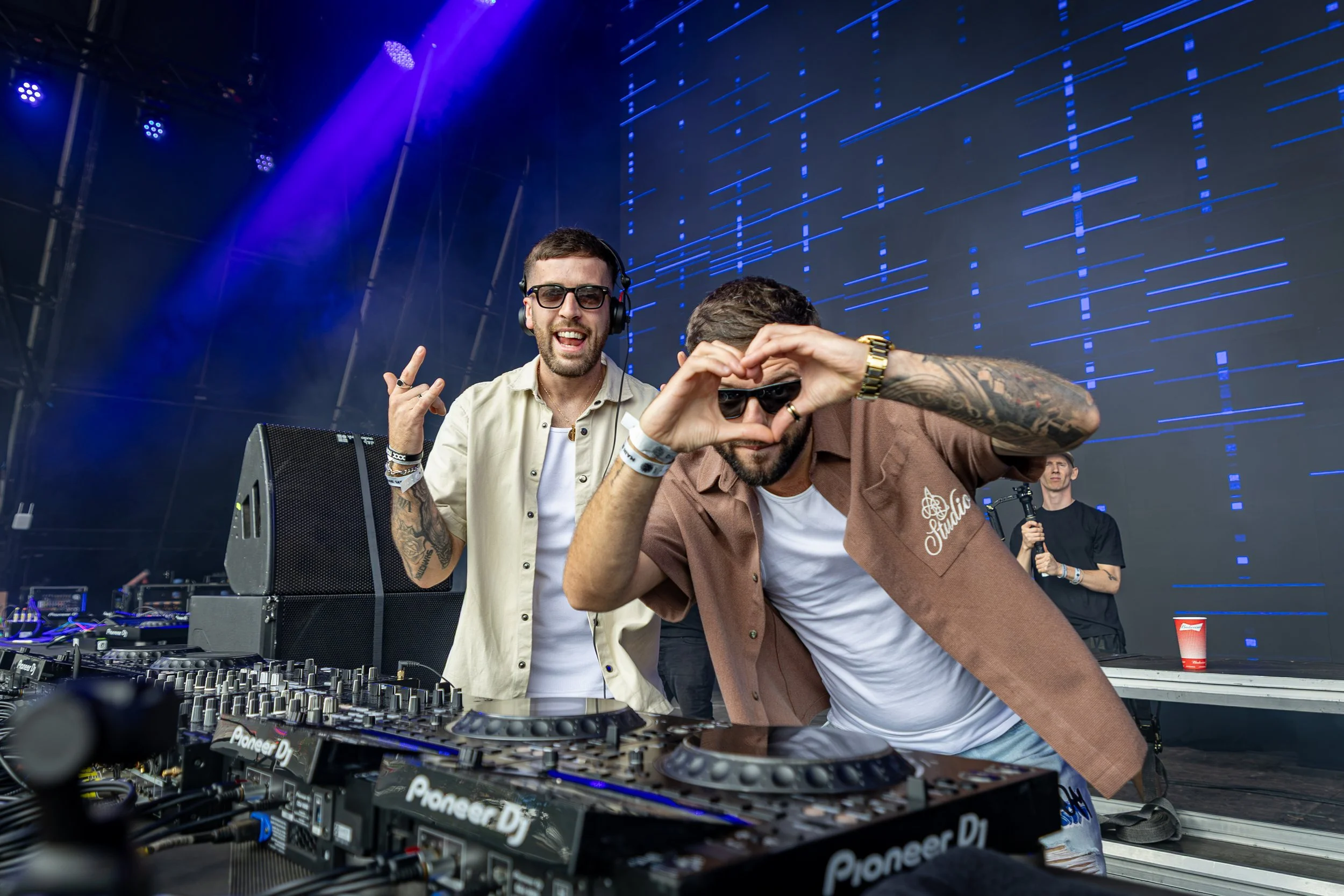 Two men DJing at an outdoor music festival with a large LED screen in the background. One man is making a heart shape with his hands while the other man is smiling and wearing headphones.