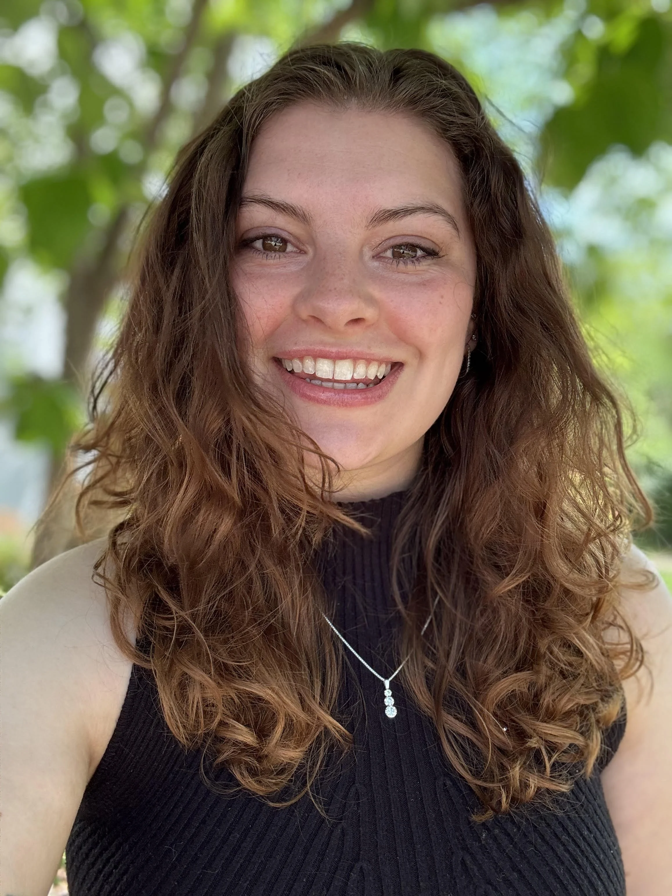 Close-up of a smiling woman with curly reddish-brown hair, wearing a black sleeveless top and a silver necklace, outdoors with green leaves in the background.
