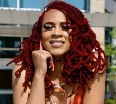 A woman with red dreadlocks smiling outdoors in front of modern buildings.