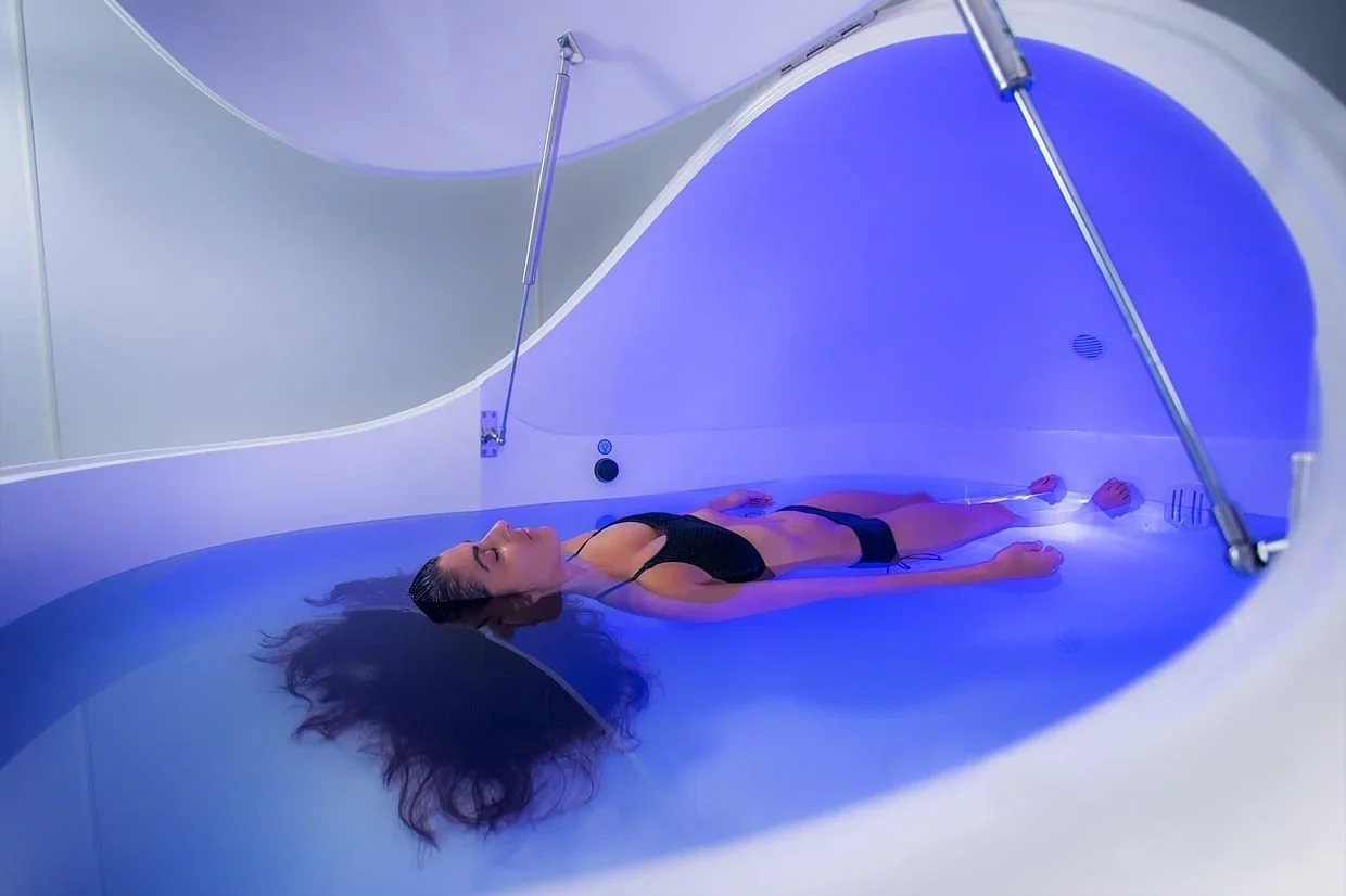 A woman lying inside a hyperbaric oxygen chamber with a transparent cover, illuminated with blue light.