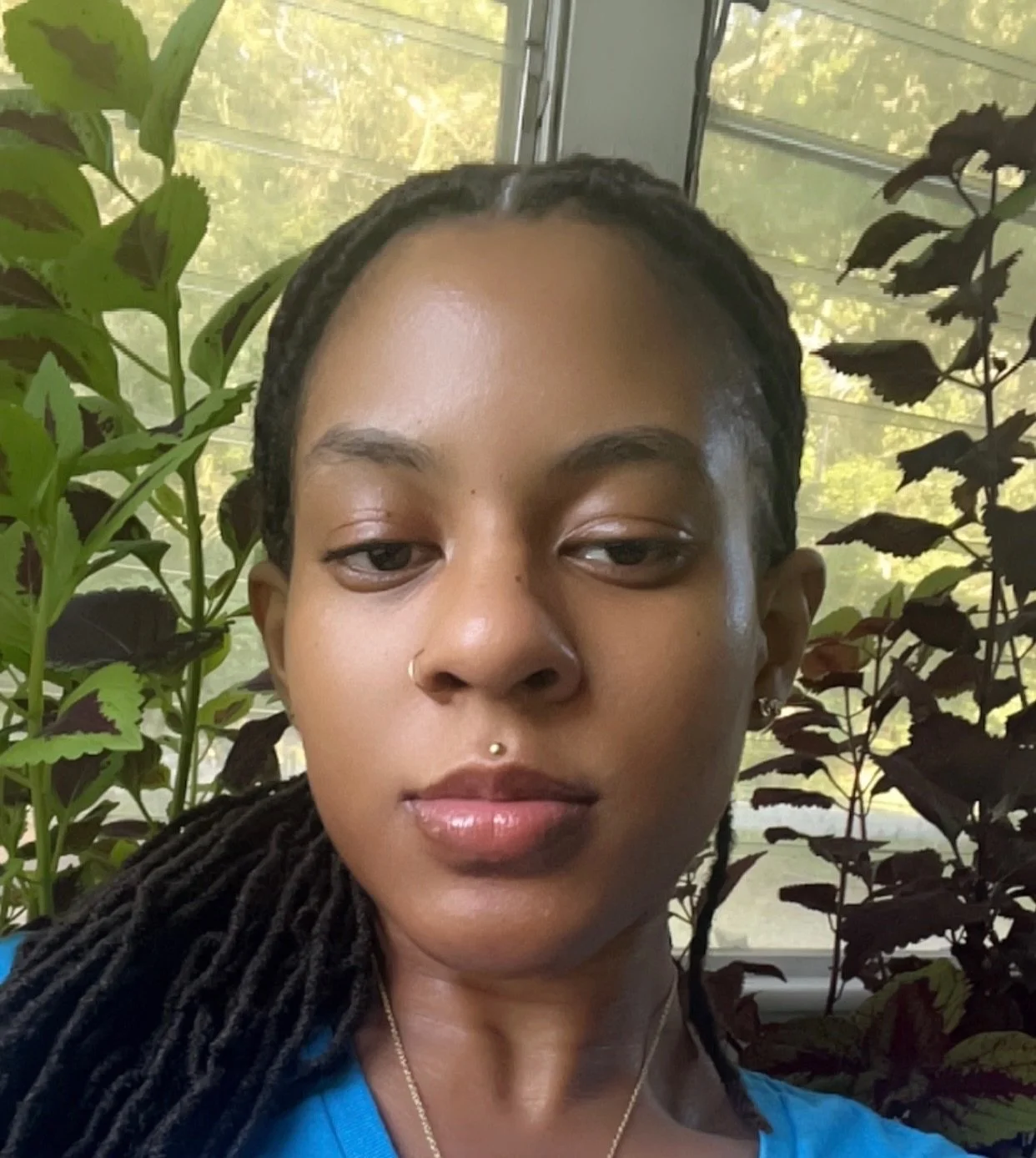 Close-up of a woman with dark braided hair and nose piercings, standing indoors near a window with green plants and trees visible outside.