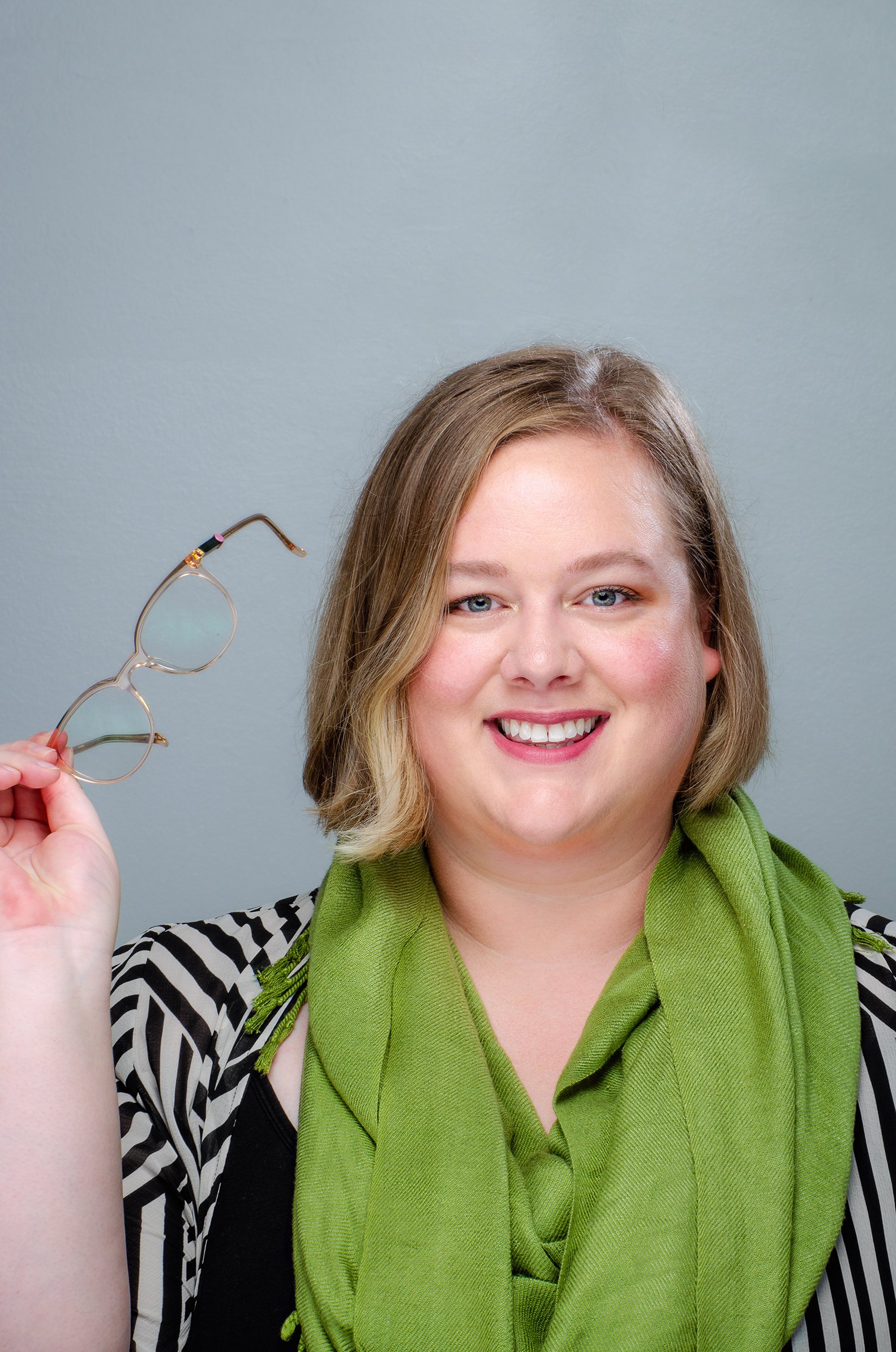A woman with blonde hair smiling while holding a pair of glasses in her right hand. She is wearing a black and white striped shirt, a green scarf, and is set against a plain gray background.