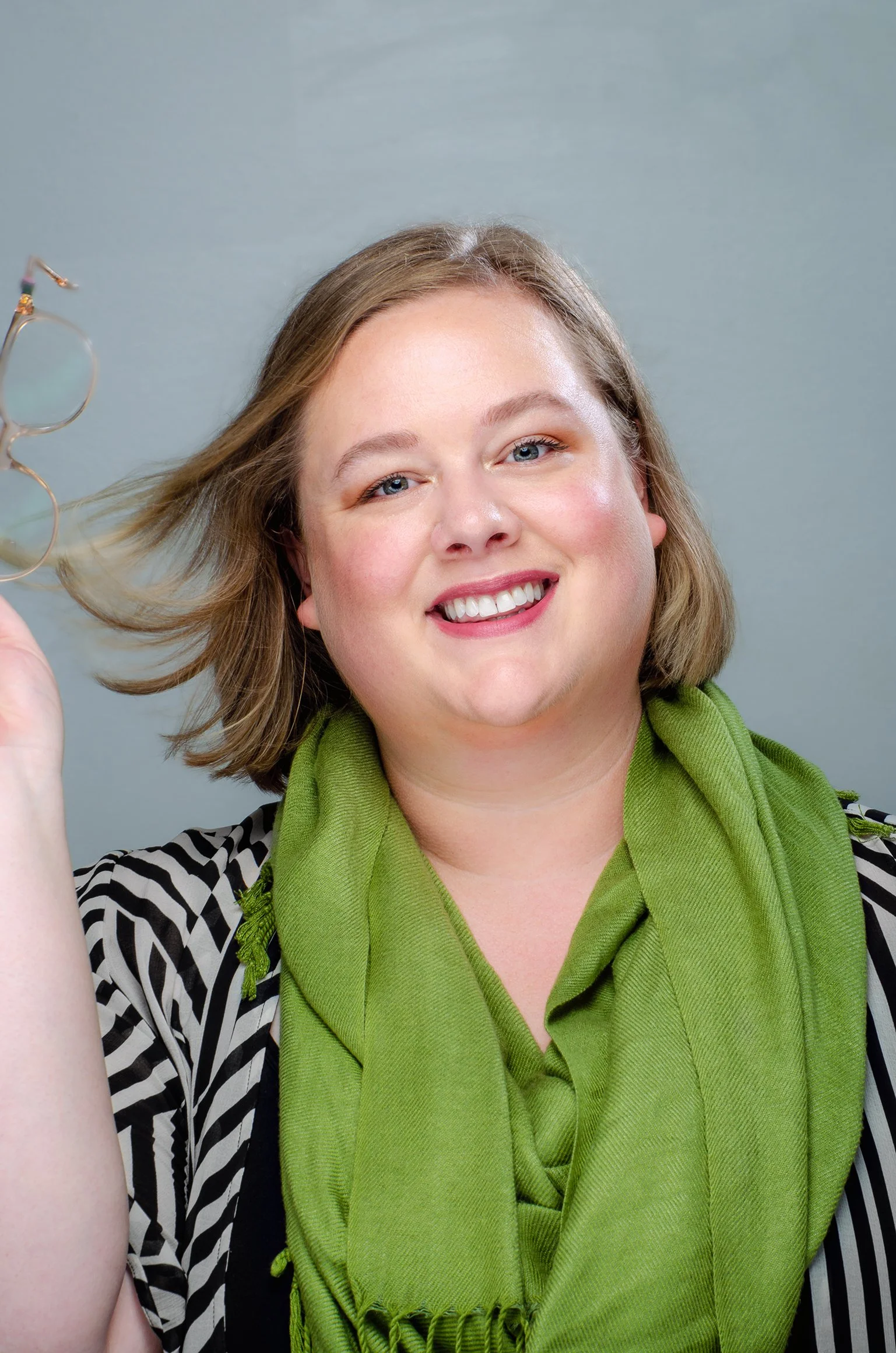 Close-up of a woman smiling, holding eyeglasses, with a gray background, wearing a green scarf and a black and white striped top.