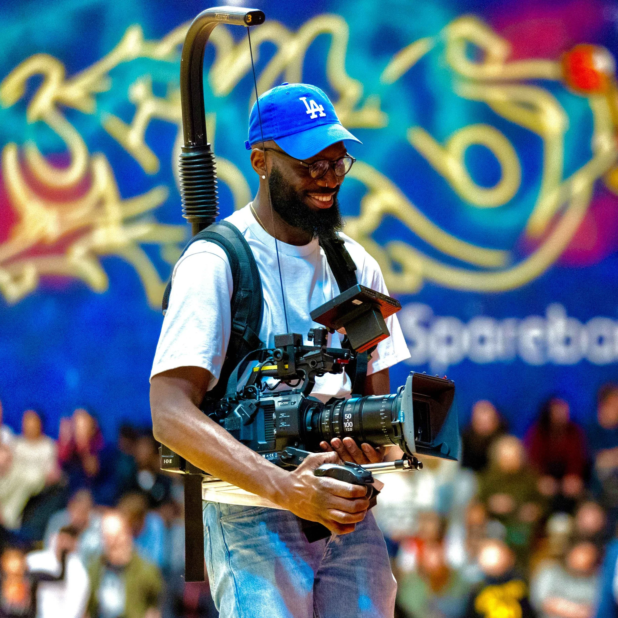 A man with a camera rig, wearing a blue baseball cap, glasses, white t-shirt, and jeans, smiling at an indoor event with a blurred crowd and a colorful mural in the background.