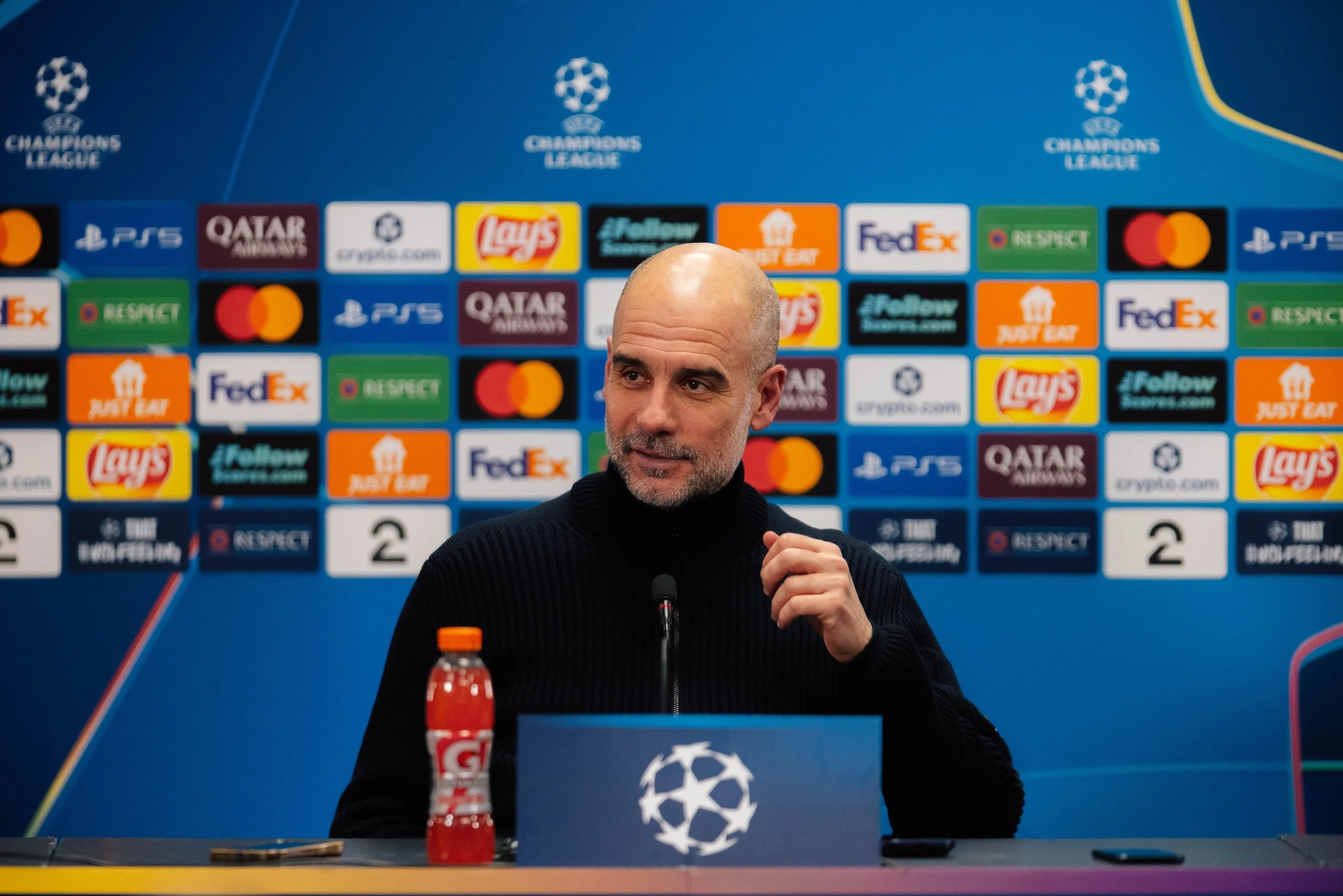 A man with a bald head and beard in a black turtleneck speaking at a UEFA Champions League press conference, sitting at a table with a microphone and a Gatorade bottle, with a backdrop featuring various sponsor logos.