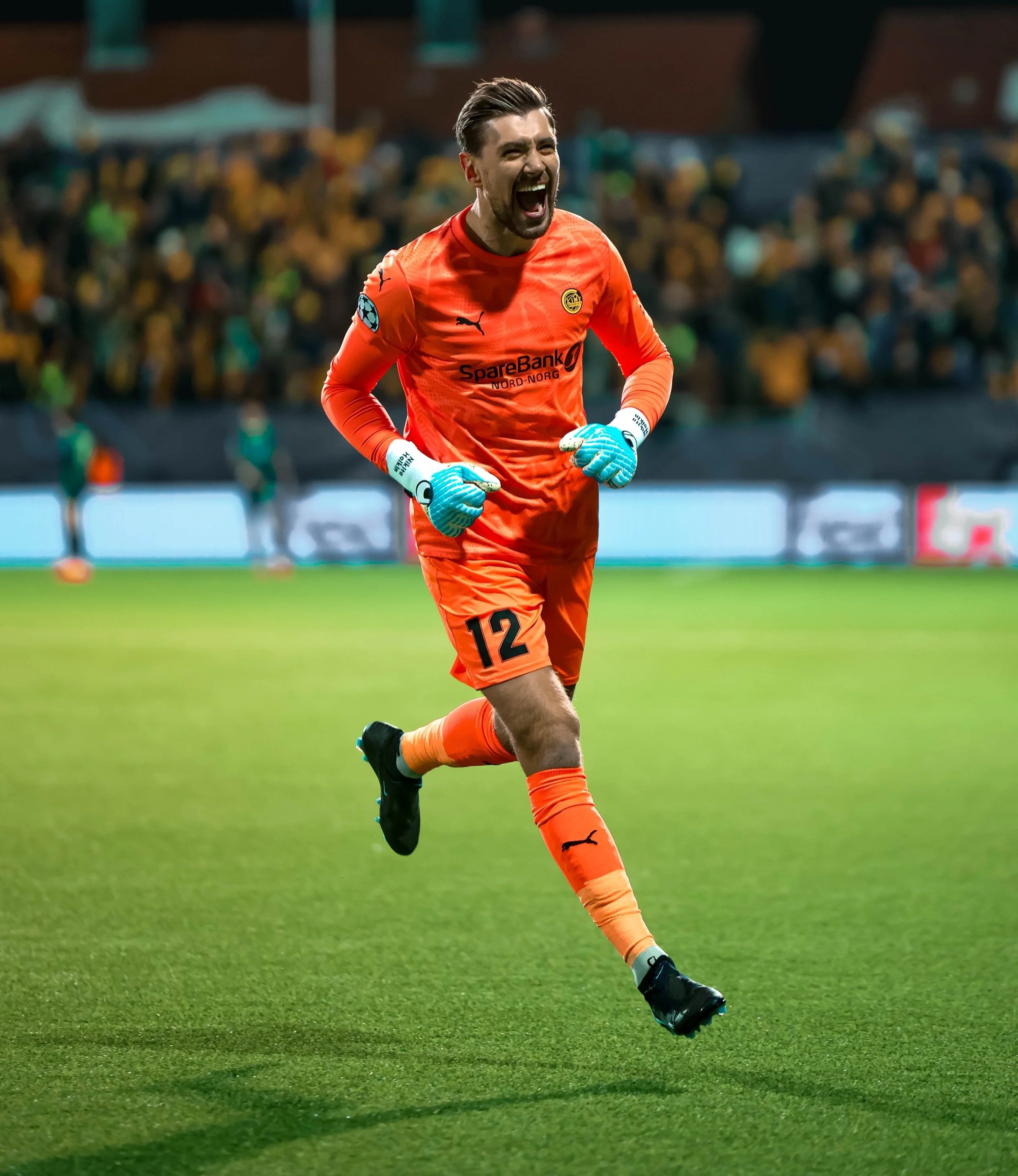 A male soccer goalkeeper in an orange uniform running on the field, celebrating with a big smile, surrounded by a crowd in the stadium.