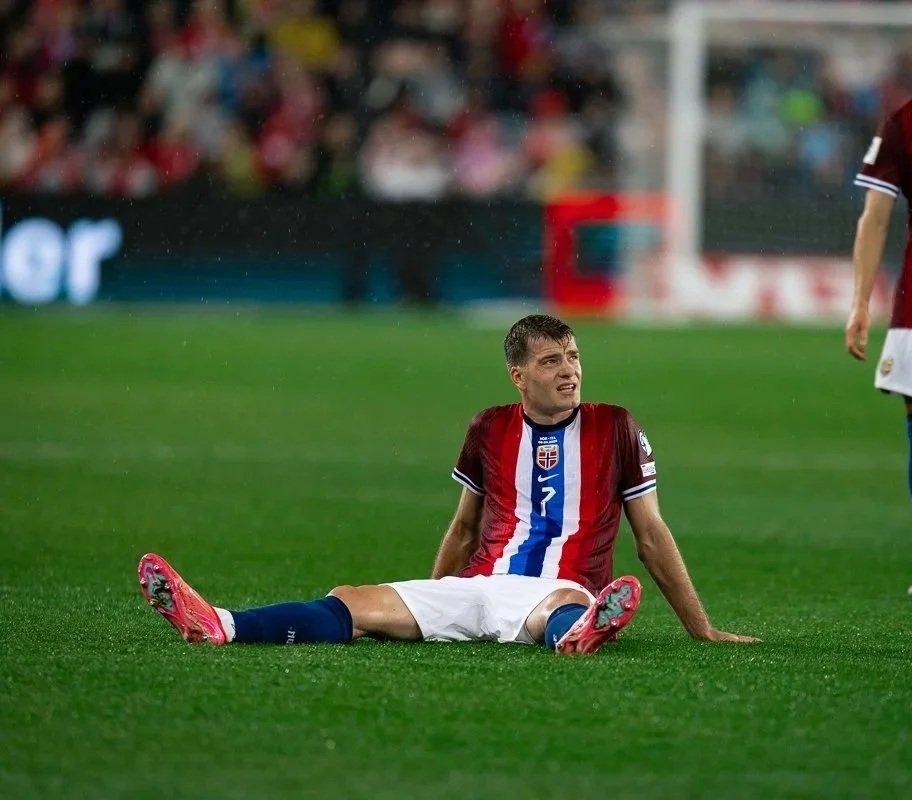 A soccer player sitting on the field with an expression of pain or frustration, wearing a red and blue jersey with a Norwegian flag emblem, pink cleats, and white shorts, while a crowd watches in the background.