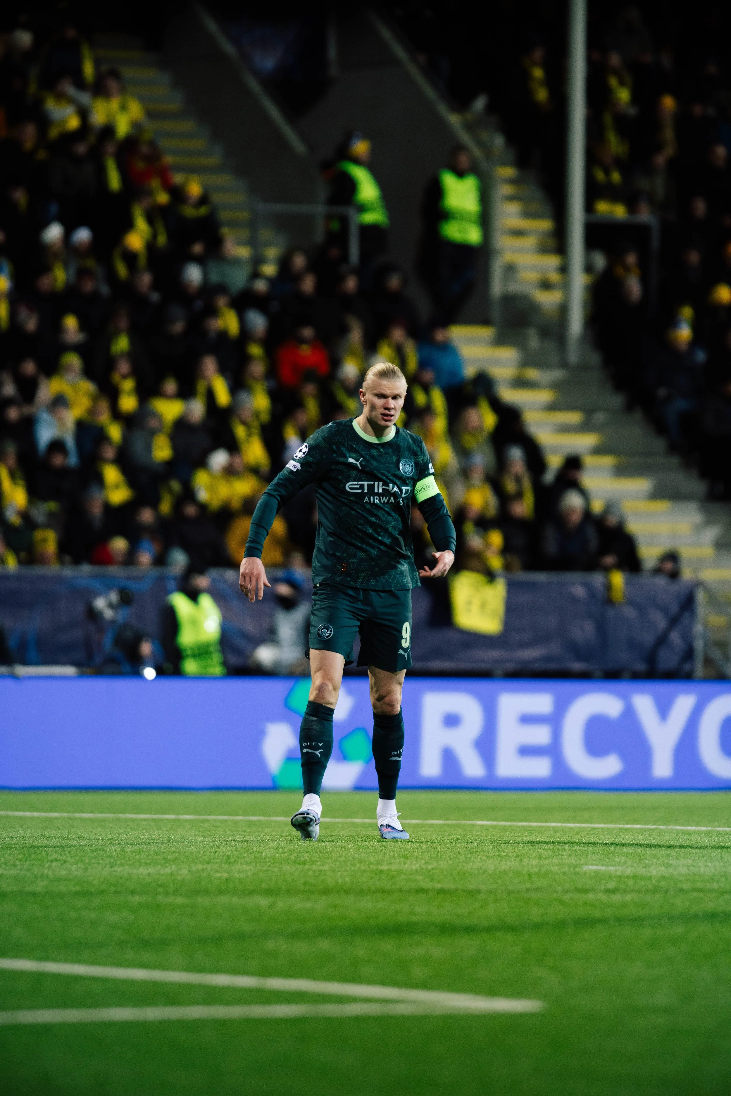 A soccer player in a dark green jersey and shorts standing on a soccer field during a game, with a crowd of spectators in the background.