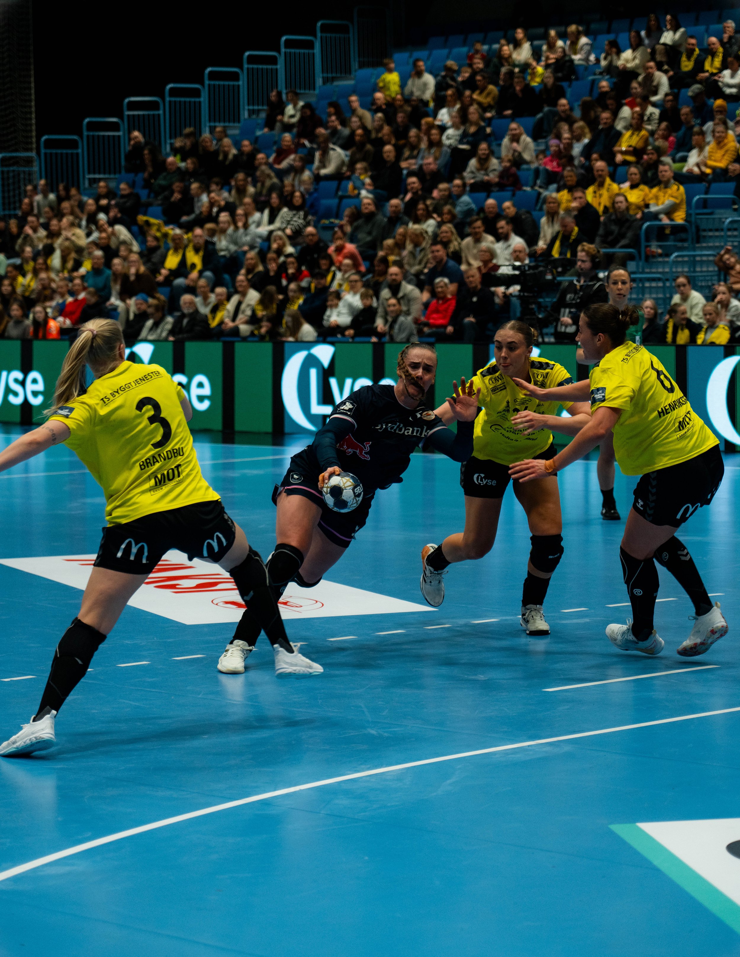A women's handball game with players in yellow and black jerseys and a black goalie on a blue court, with spectators in the background.