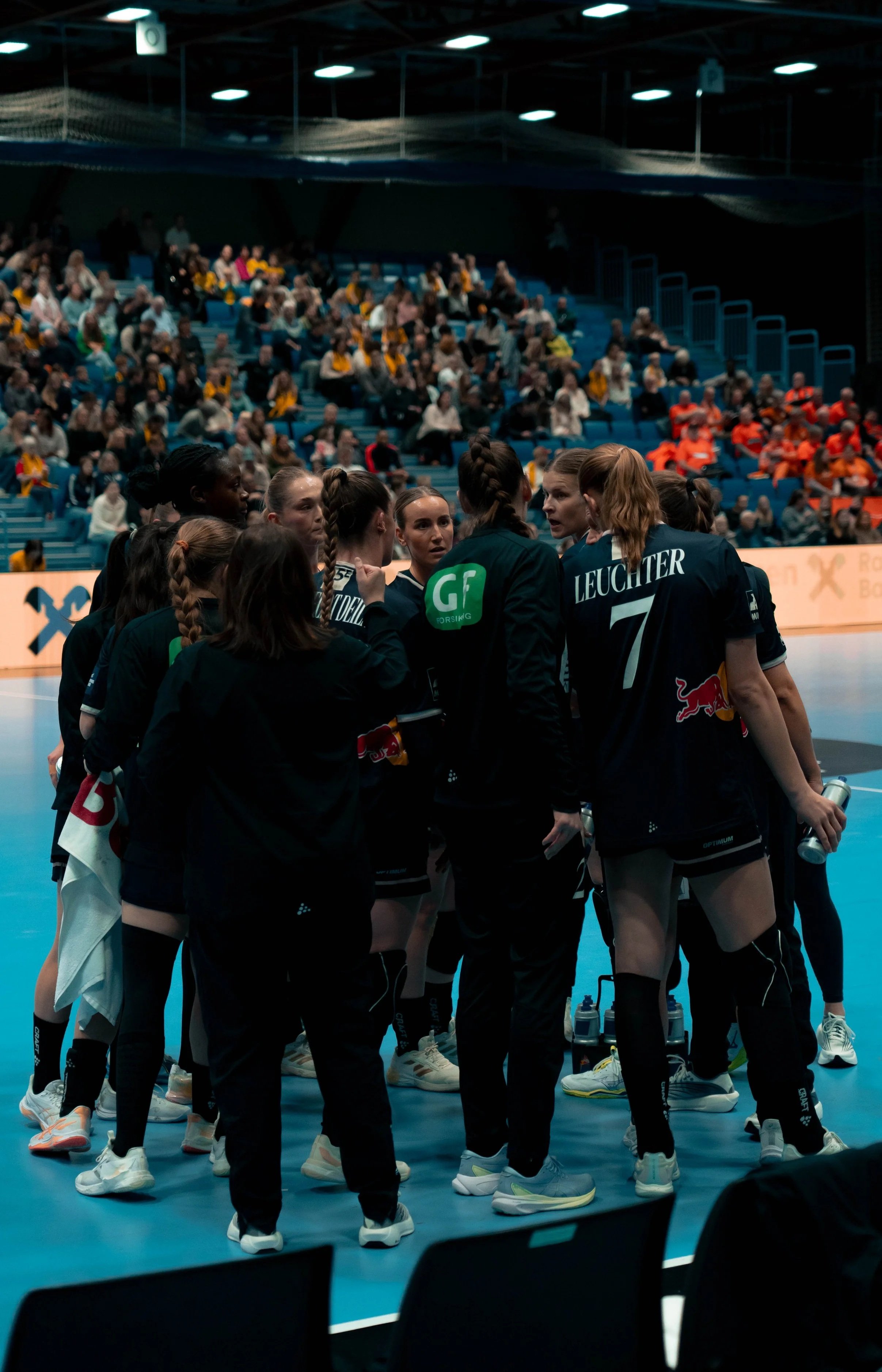A group of female athletes in black uniforms with sponsor logos, gathered in a huddle on an indoor sports court during a game, with a crowd of spectators seated in the background.