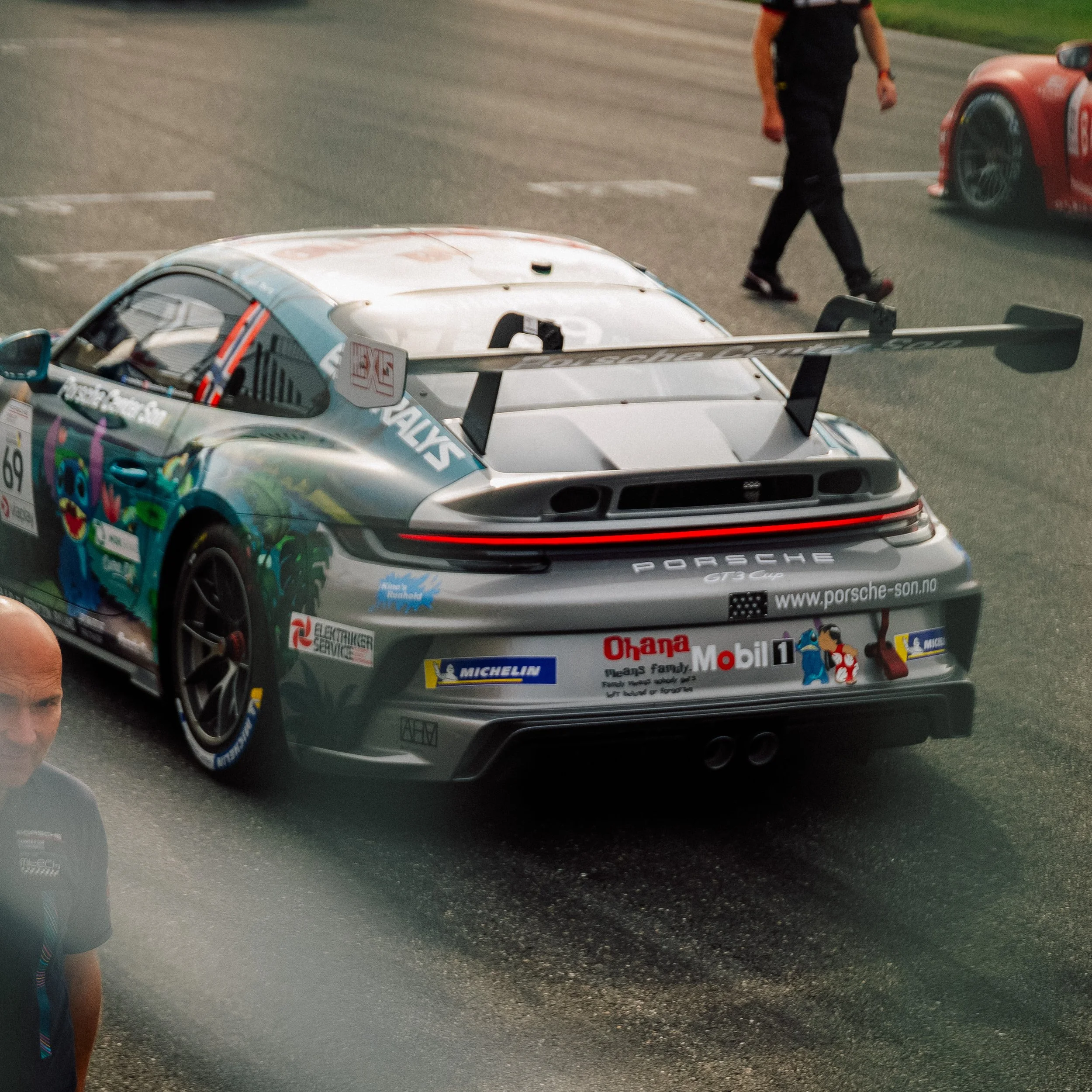 Photo of a silver Porsche race car on a track, with sponsor stickers and a large rear wing, surrounded by team members.