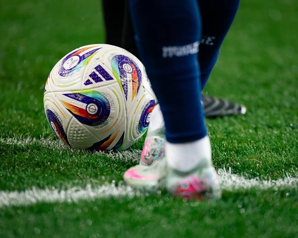 Close-up of a soccer ball on the grass with a player preparing to kick it, showing the player's legs and shoes.