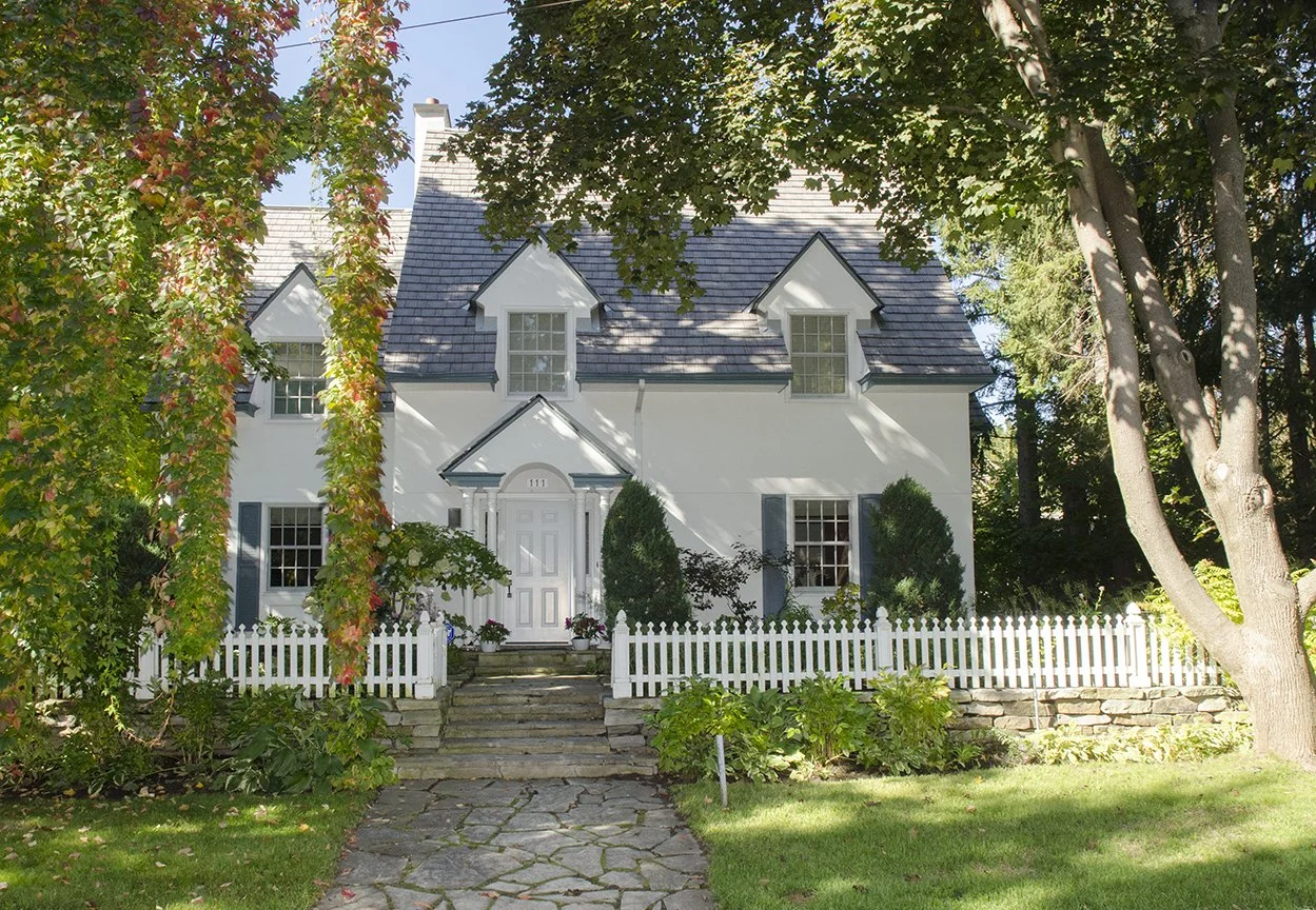 A white two-story house with a steep gray roof, surrounded by trees, a white picket fence, and a stone walkway leading to the front door.