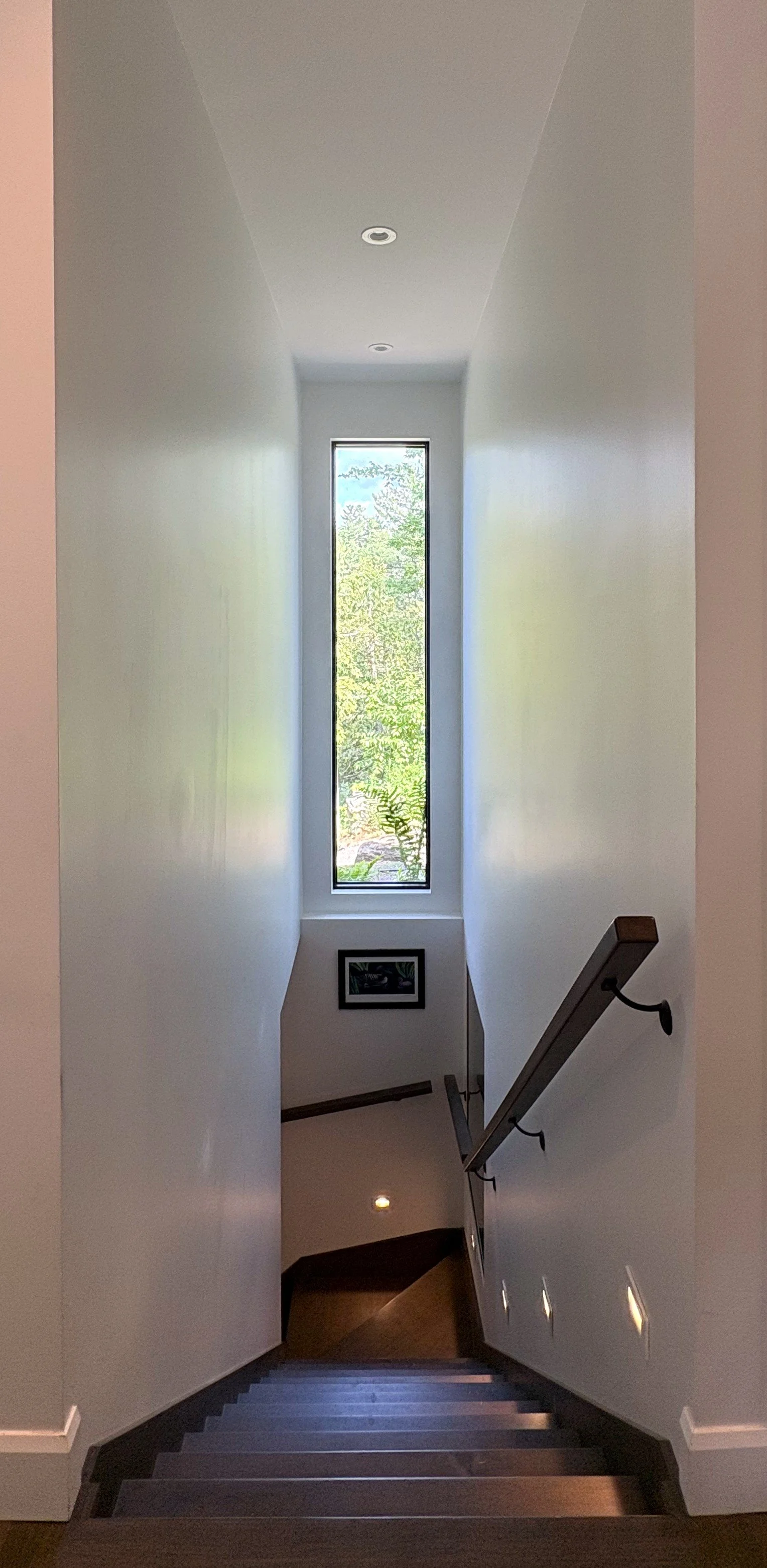 Interior of a staircase with wooden steps, a simple handrail, a large vertical window letting in natural light, and a framed picture on the wall at the top of the stairs.