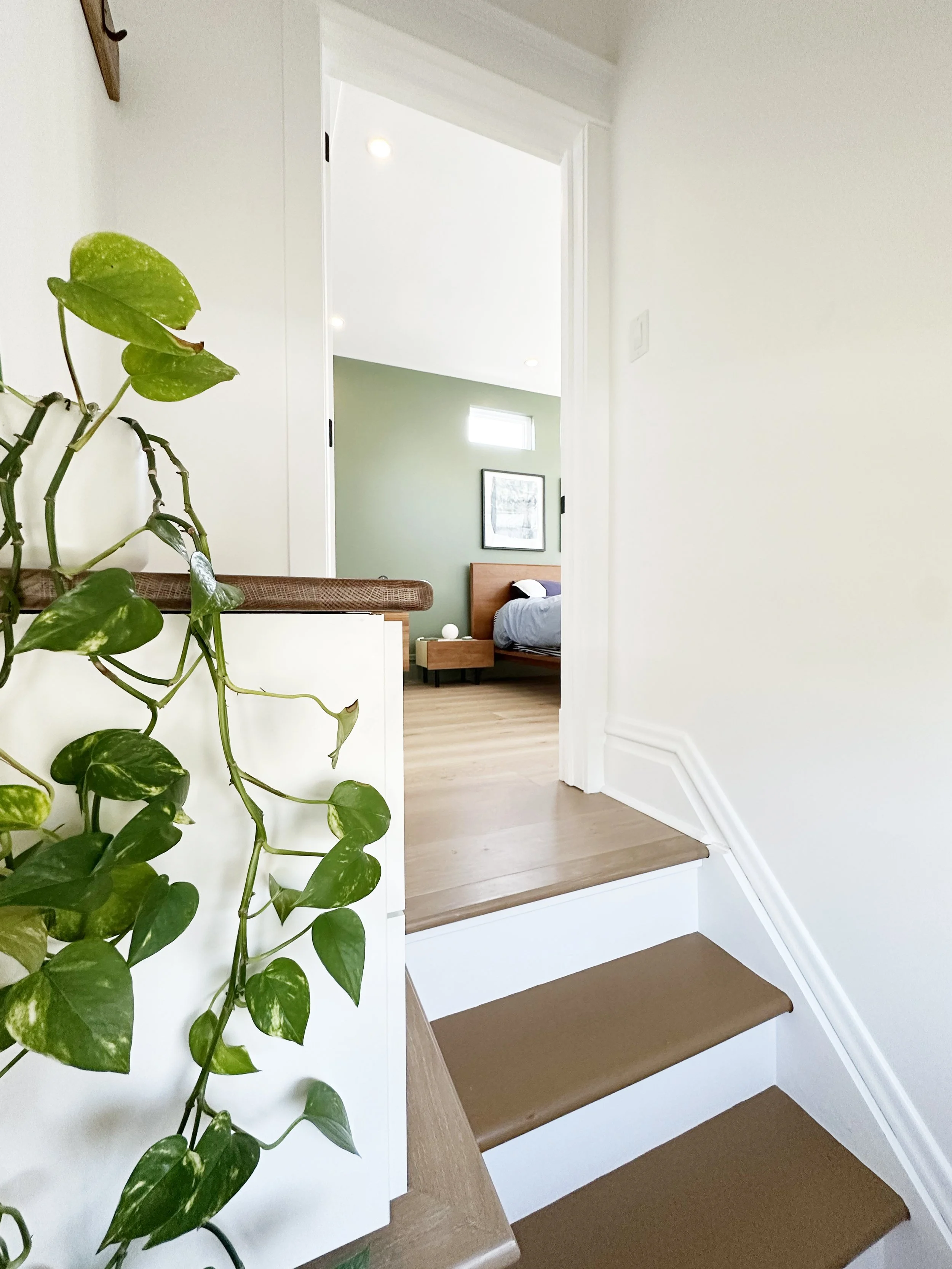 View of a staircase with wooden steps, white trim, leading to a bedroom with a bed, nightstand, and artwork on the wall, as seen through an open door, with a green accent wall and a window letting in natural light.