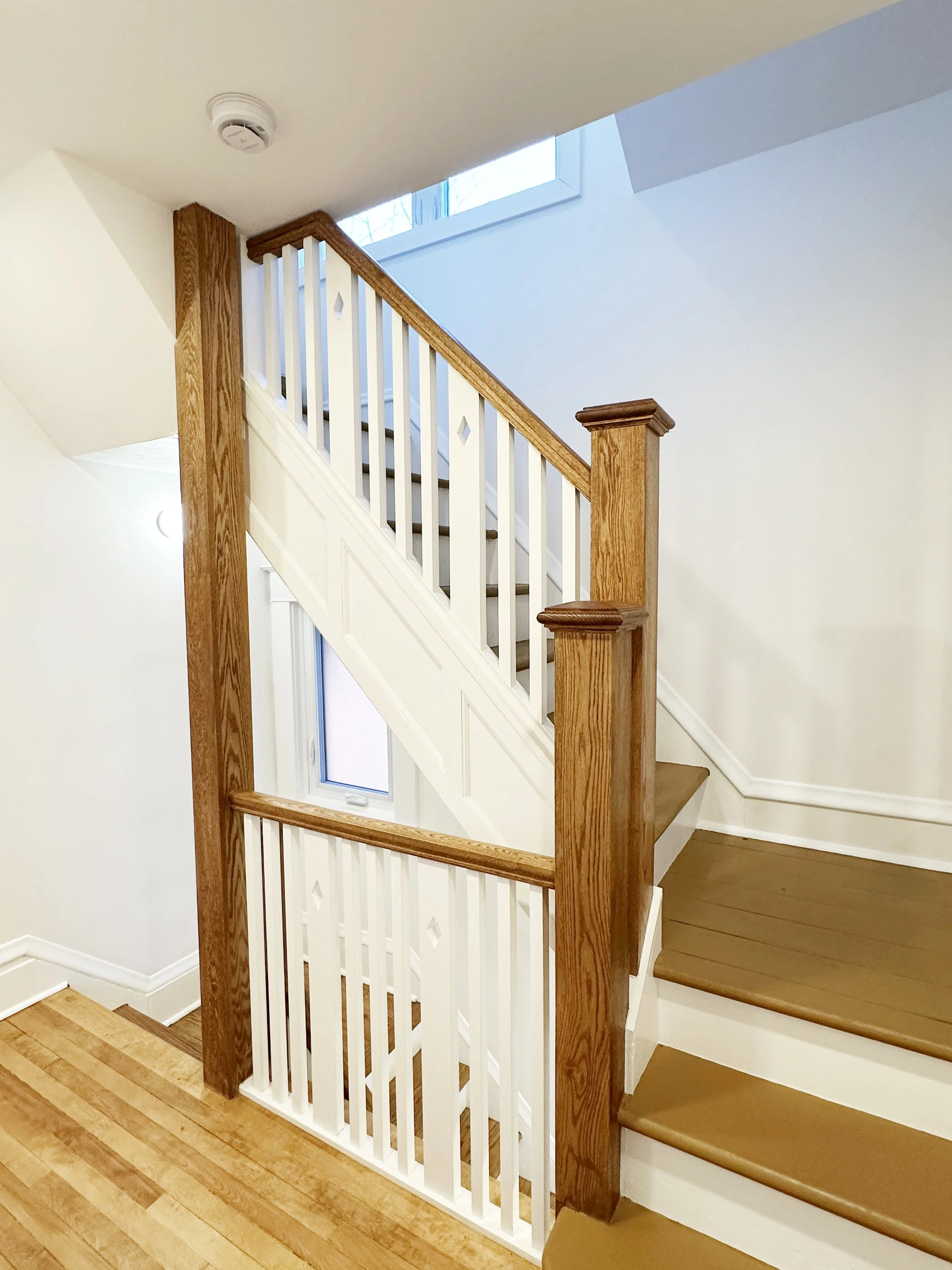 Interior view of a staircase with wooden handrails and white balusters, adjacent to a wall with a window allowing natural light to illuminate the area, and hardwood flooring.