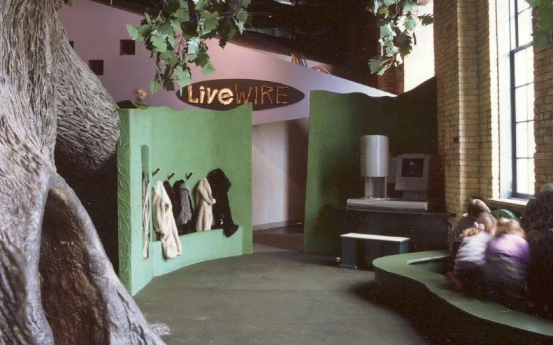 Indoor space with green walls, coat hooks with hanging clothes, a large artificial tree trunk, and a group of children sitting on a curved bench near a window.